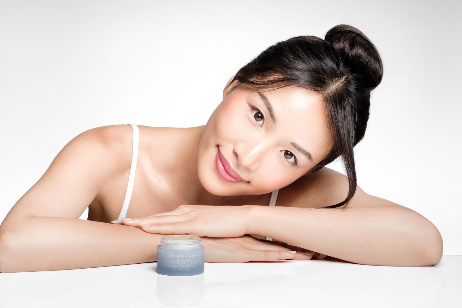 Young Asian woman with hair bun smiling beside a Summer Fridays moisturizer cream jar on a white surface.