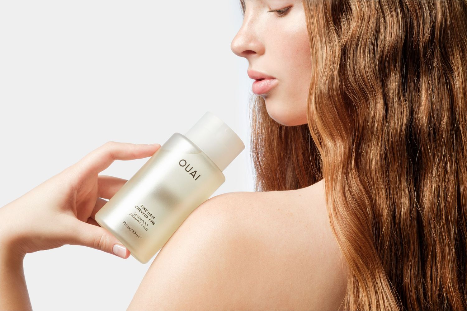 Woman with long wavy hair holding an OUAI fine hair shampoo bottle over her shoulder against a light grey background.