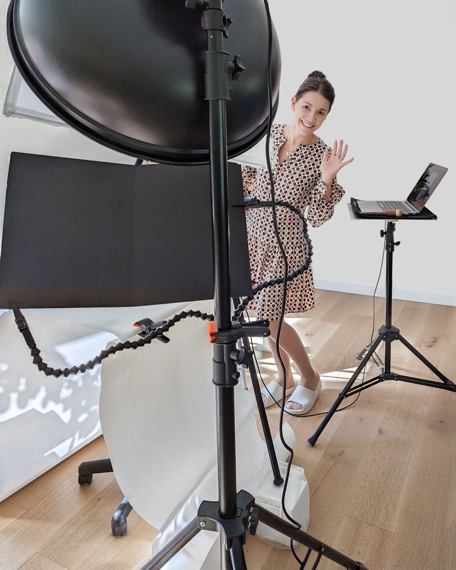 Darina in her home studio waving at the camera positioned in the middle of her setup with lighting equipment and a laptop on a stand.