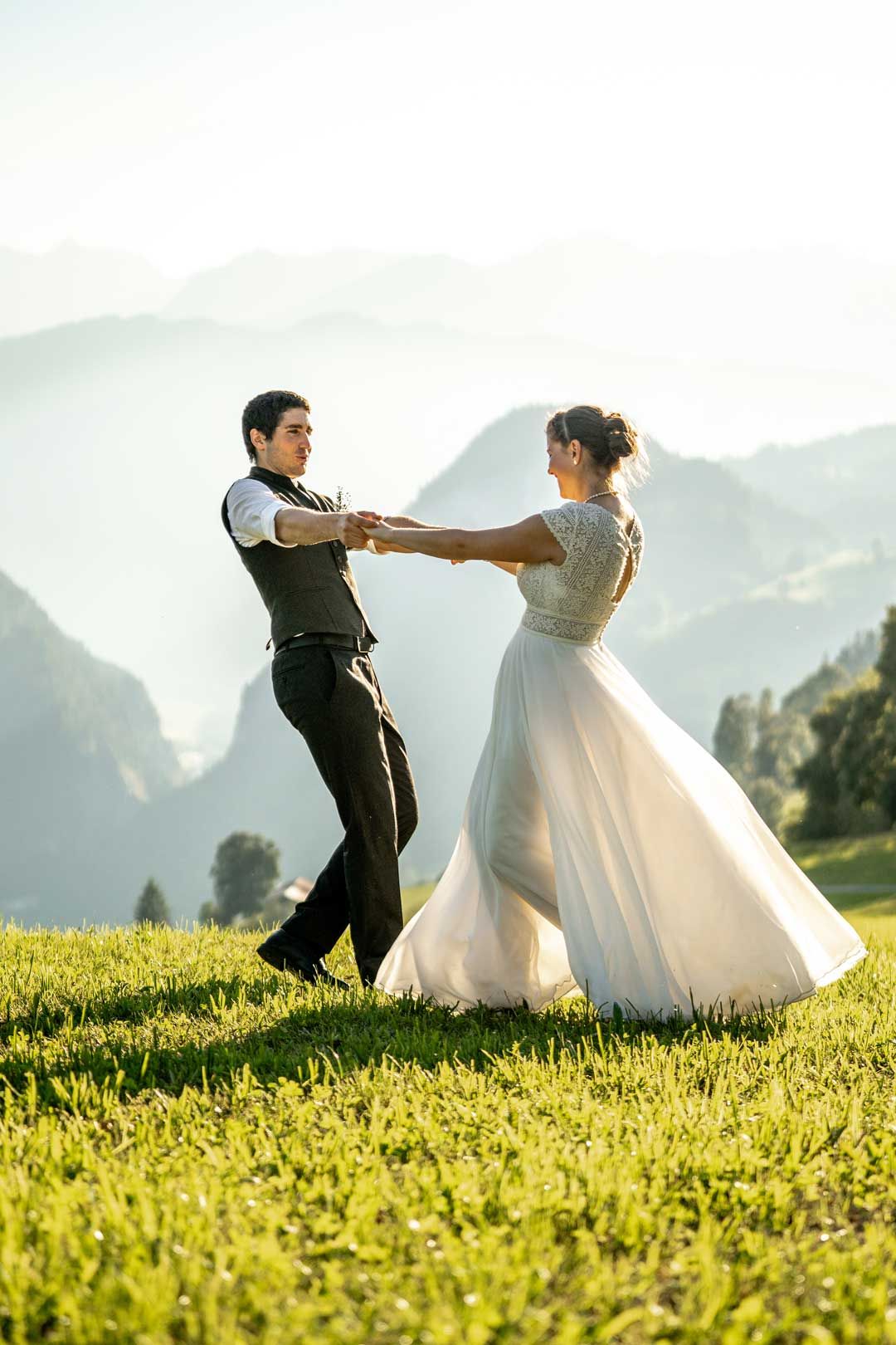 Hochzeitsfotografie: Hochzeitspaar tanzt hoch in den Bergen auf einer Wiese – beide strahlen überglücklich