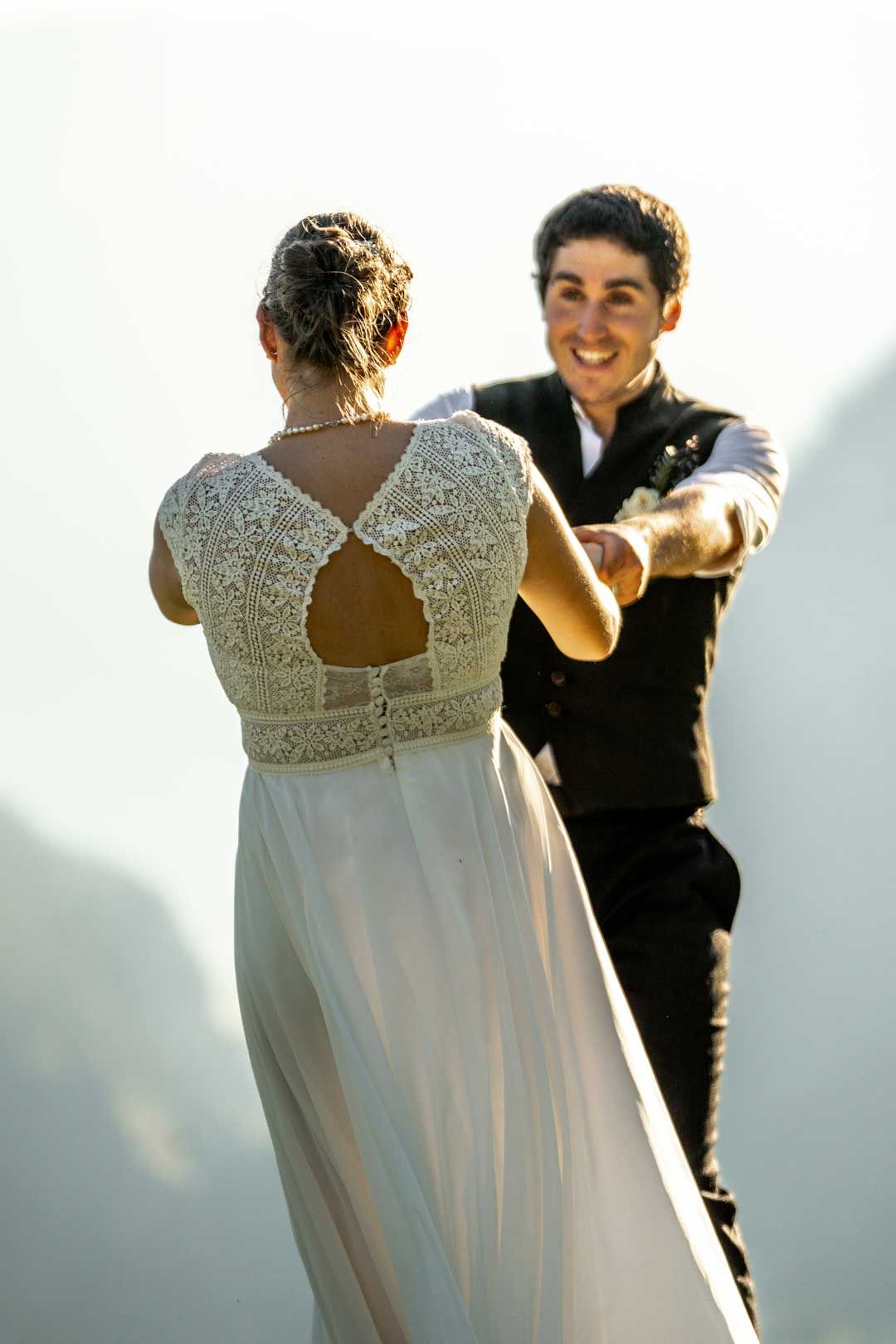 Hochzeitsfotografie: Hochzeitspaar tanzt hoch in den Bergen auf einer Wiese – beide strahlen überglücklich