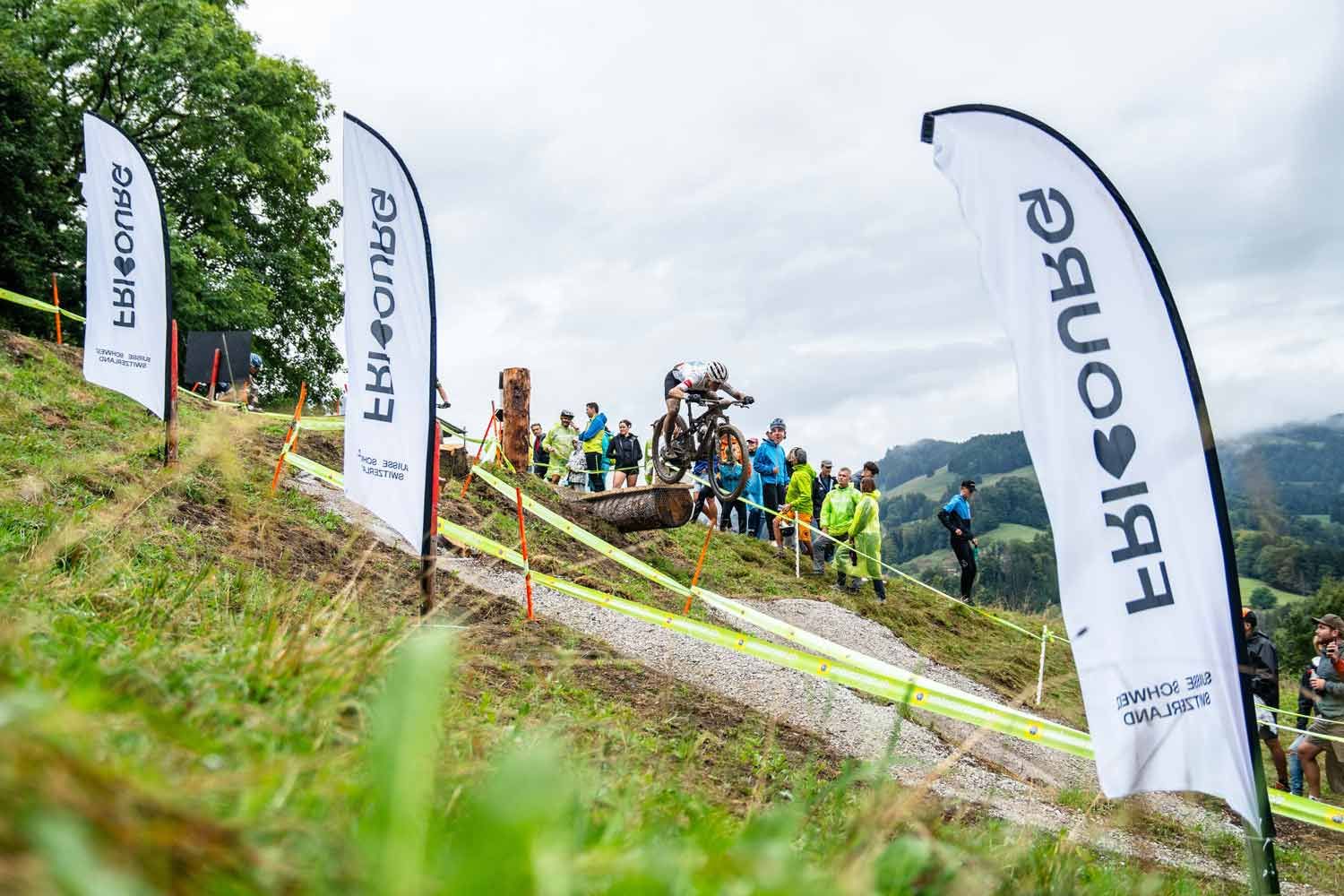 Eventfotografie: Ökk Bike Revolution in Gruyere – Mountainbikerin fährt über Drop und wird angefeuert von Zuschauern