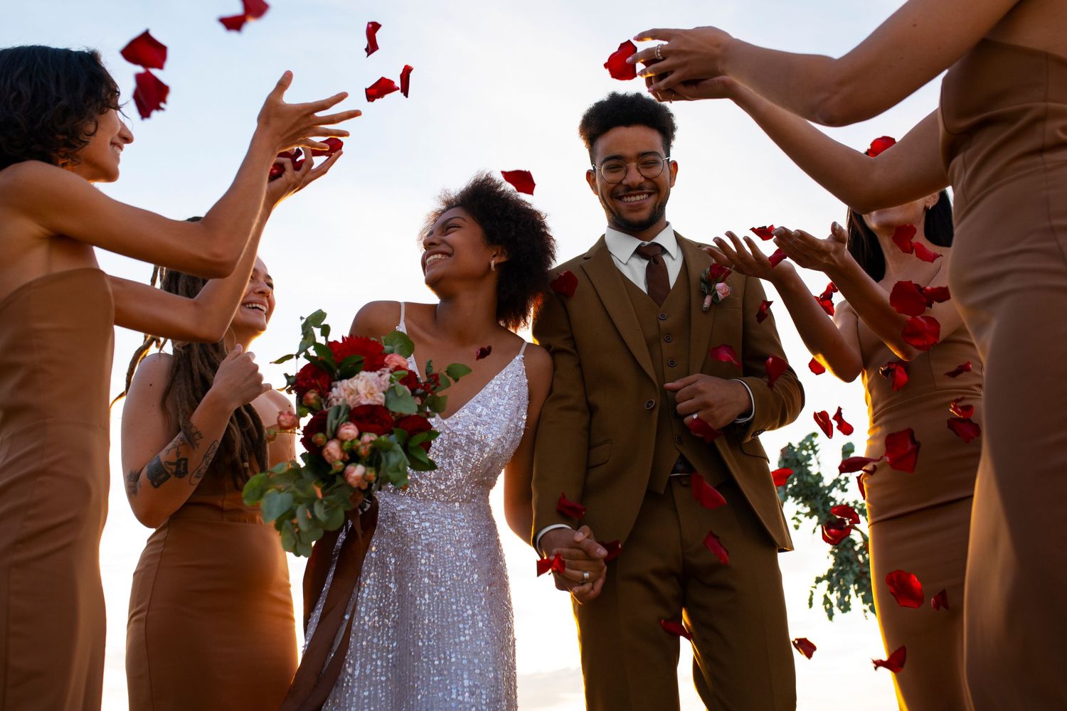 Red rose petals fall between a bride and groom during an outdoor wedding celebration.