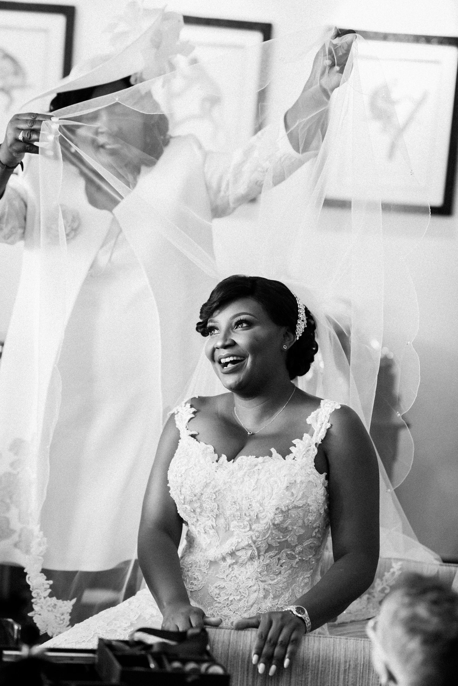 A joyful bride in a lace gown laughs while someone adjusts her flowing veil in a black and white photo.