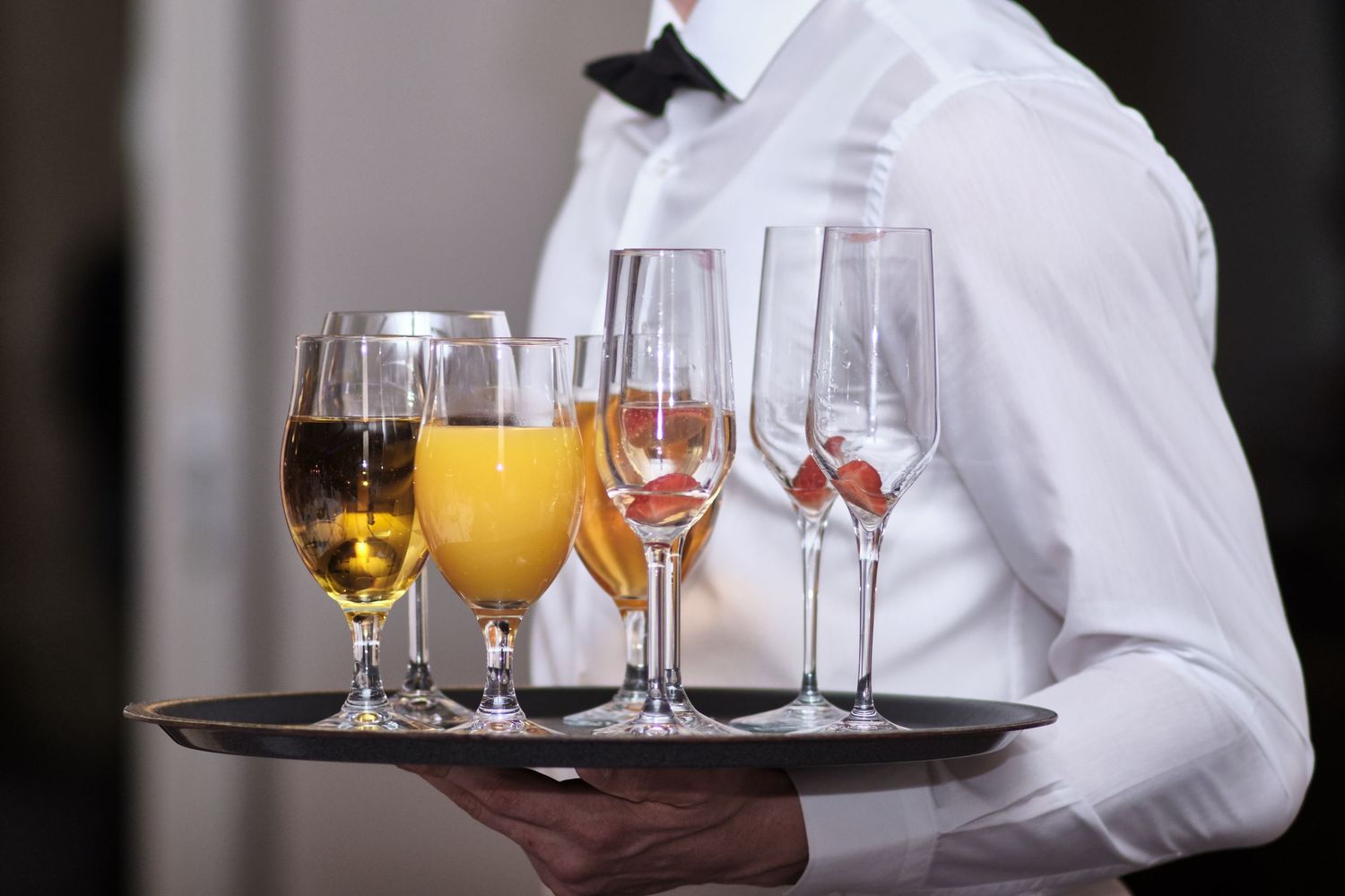 Professional server in white uniform carries a tray with assorted beverages and glassware in an upscale setting.