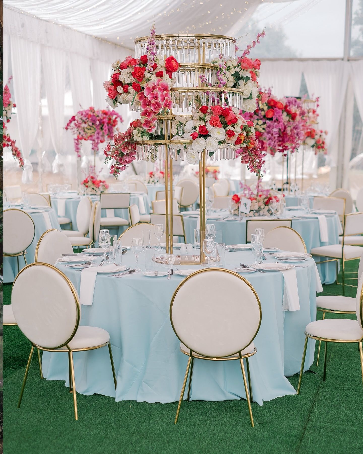 Elegant wedding reception tables with pink floral centerpieces, gold chandeliers, and white chairs on artificial grass.