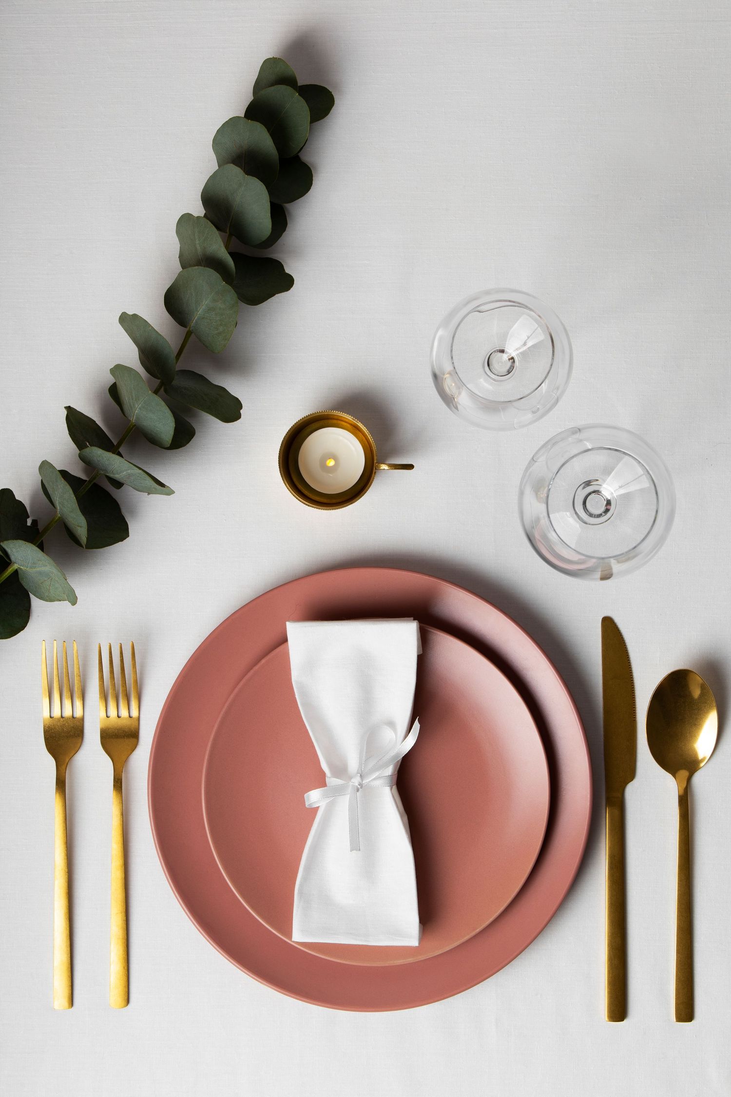 Elegant table setting with dusty pink plates, gold cutlery, wine glasses and eucalyptus branches on white background.