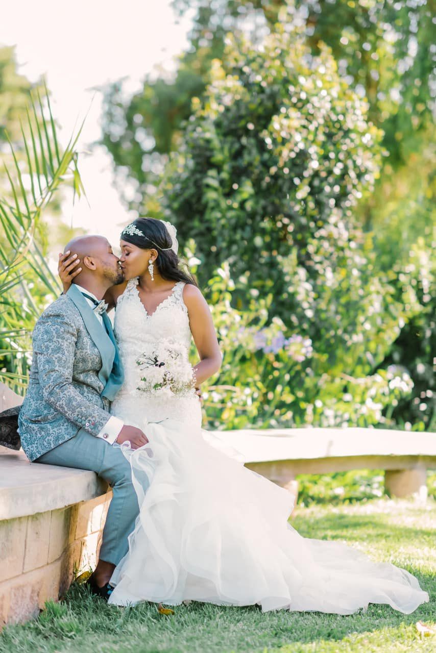 A romantic wedding couple shares intimate moments while sitting on a stone bench in a lush garden setting.