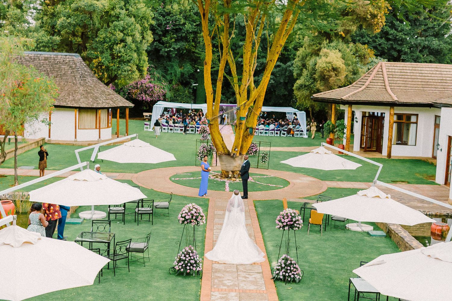 Outdoor wedding venue setup with white tents, green lawn, and rustic decor in a garden setting during daytime.