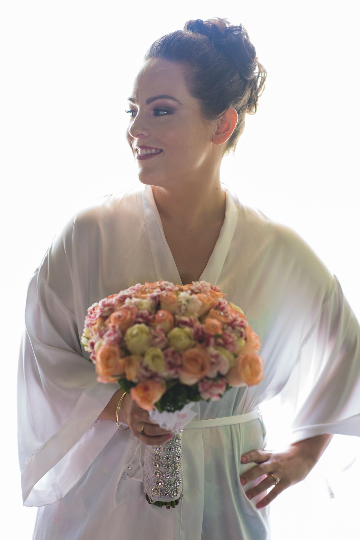 Three bridal party members in white robes smile and pose with peach-colored floral bouquets in a bright setting.