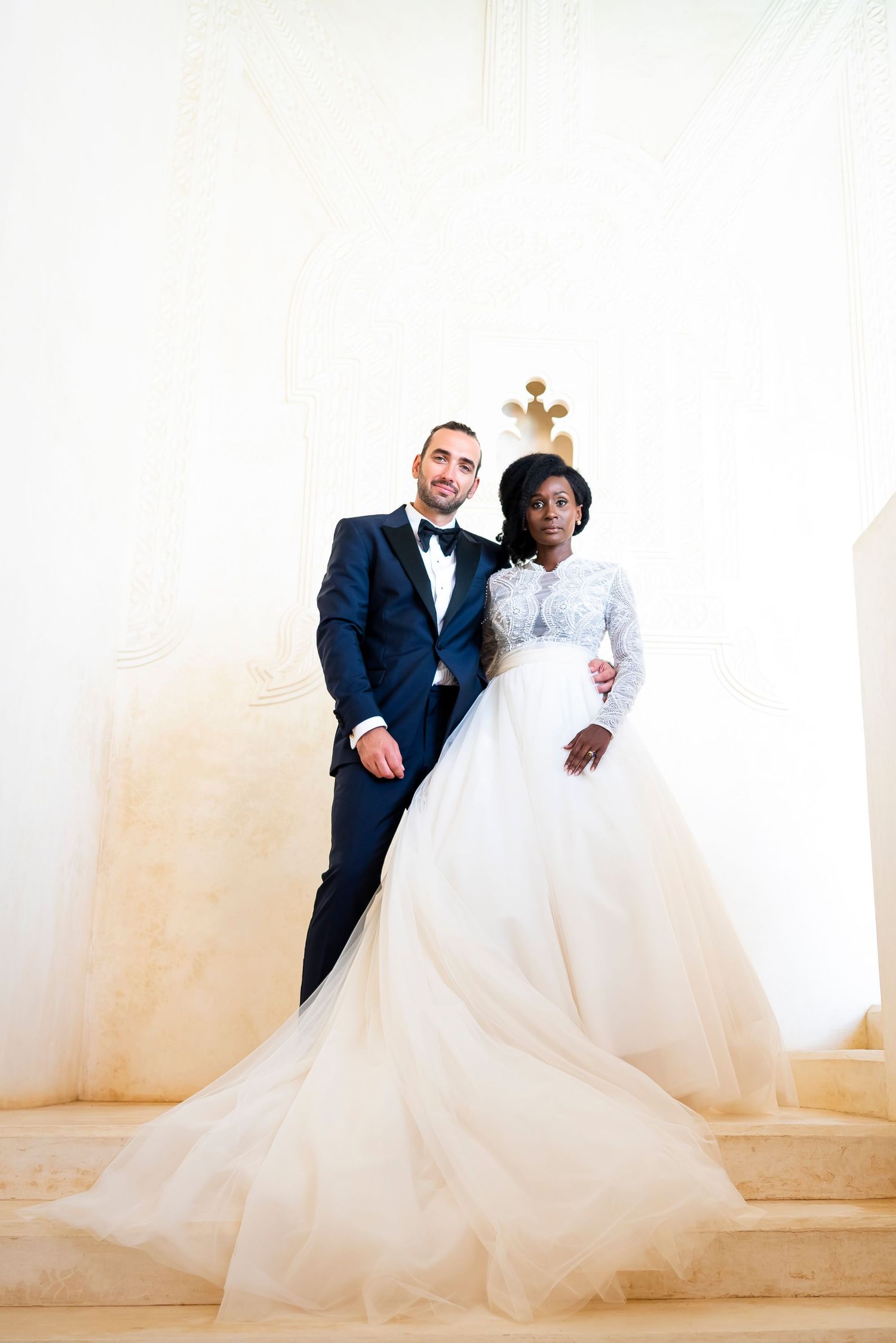 A bride and groom pose together against a light colored wall in an elegant wedding portrait.