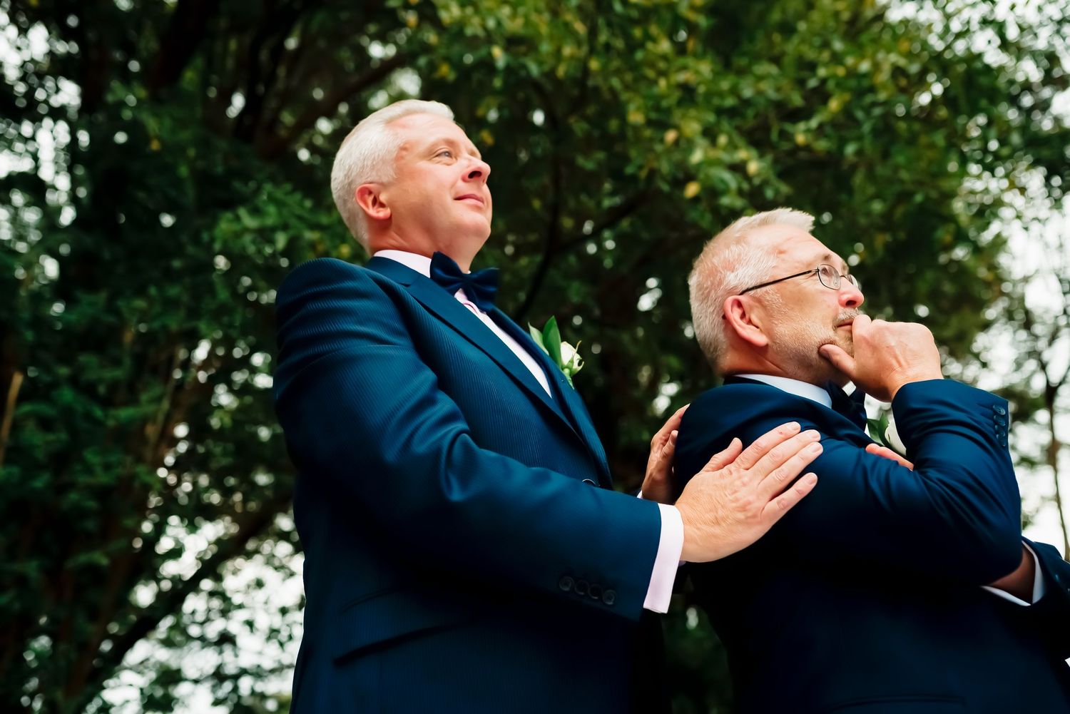 Two senior men in navy suits react emotionally while standing outdoors with trees in the background.