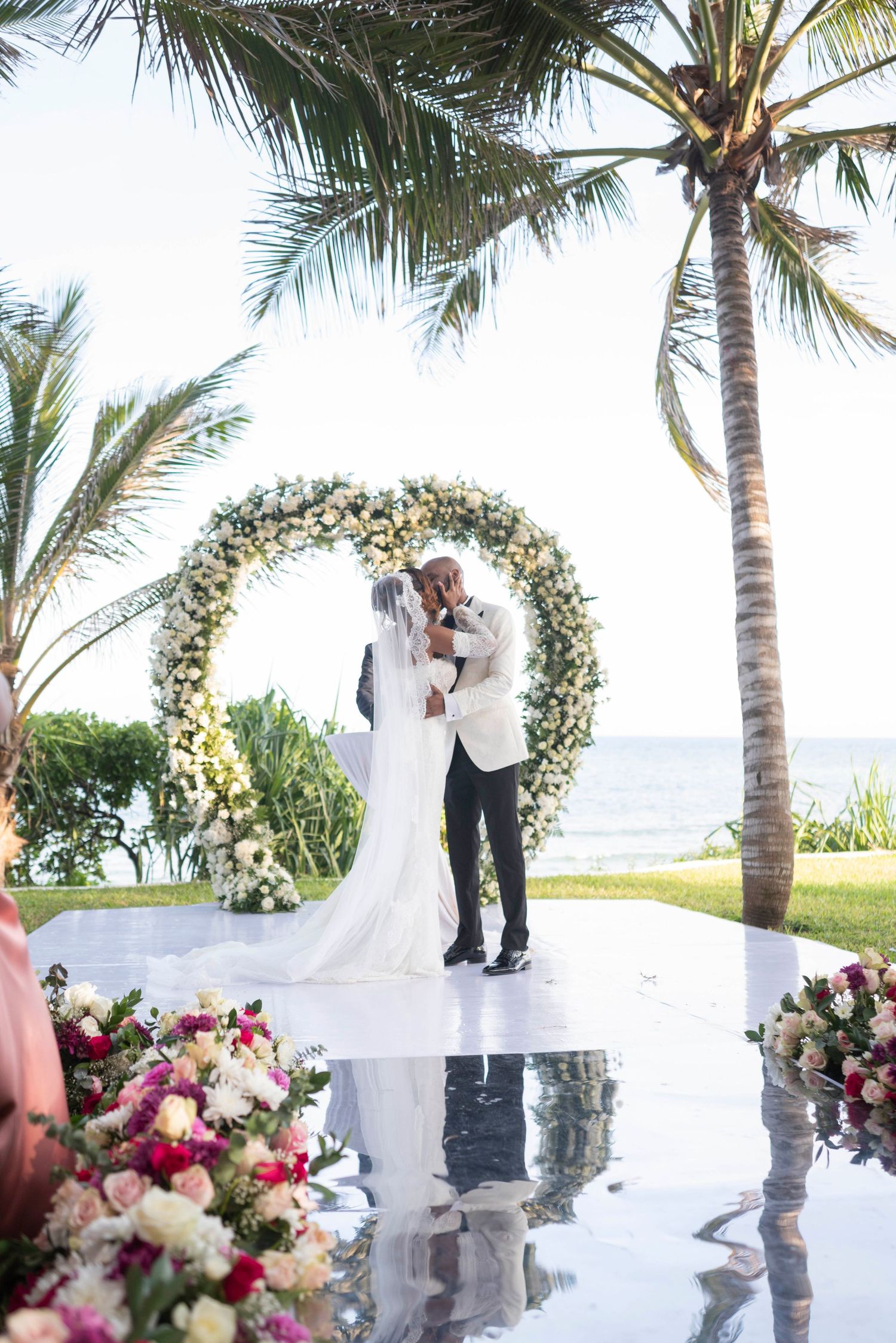 Romantic outdoor beach wedding ceremony under palm trees with white floral arch and ocean view.