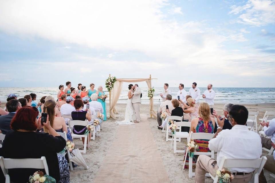 A panoramic view of a beach wedding ceremony with white chairs and guests gathered under a sunny blue sky.