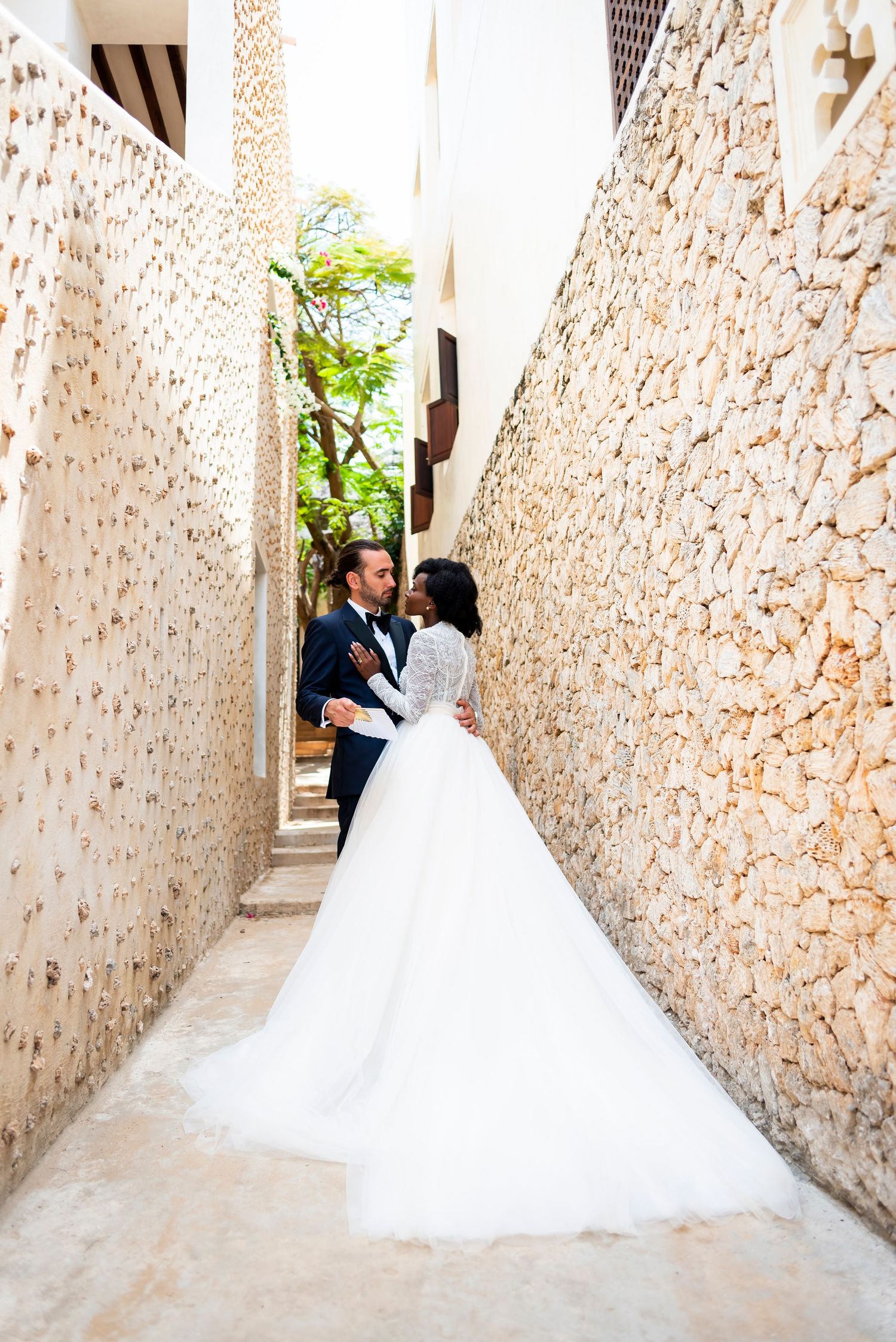 Newlywed couple walking down a rustic stone-walled corridor with a flowing white wedding dress trailing behind them.