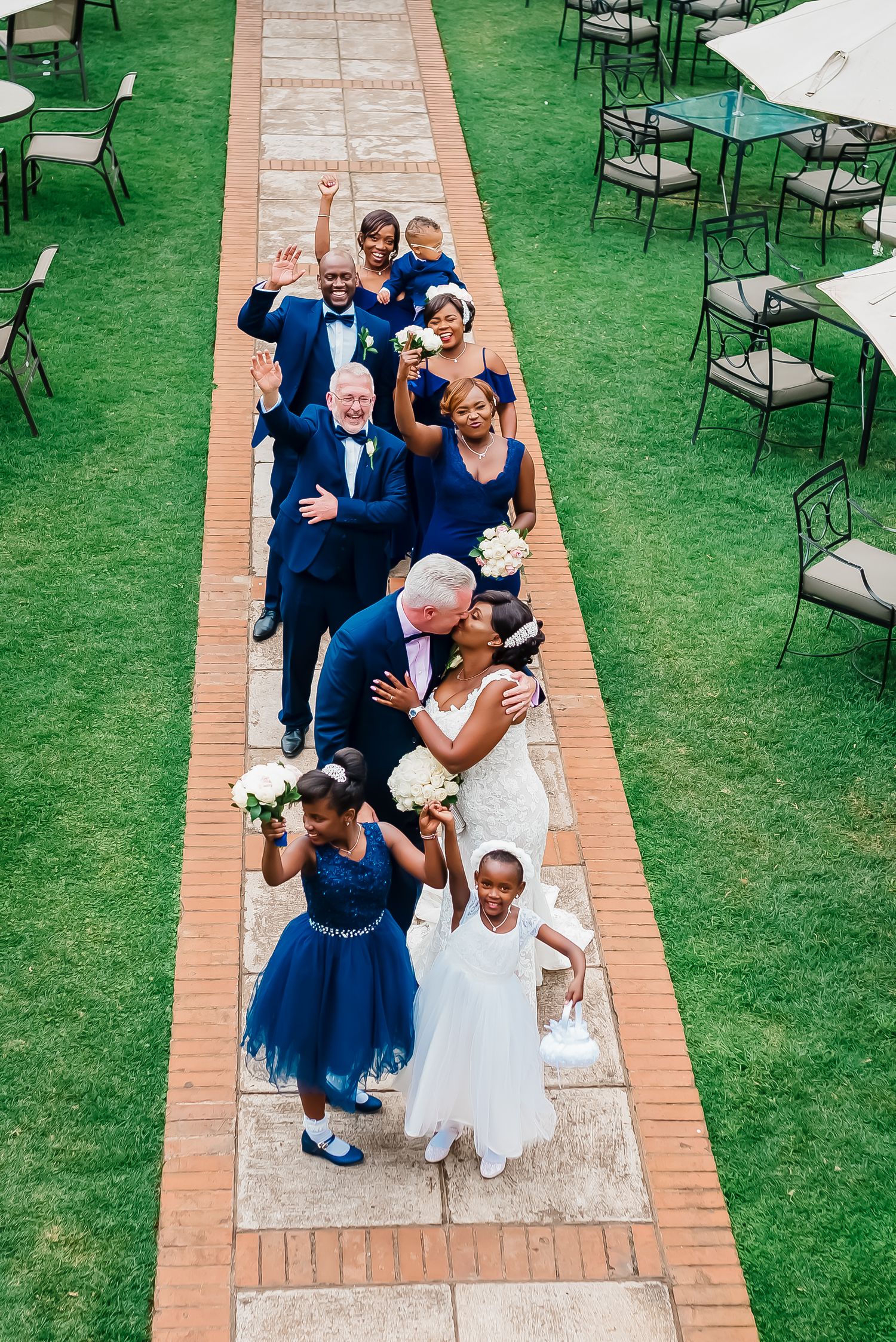 Wedding party poses in a line on brick pathway with blue dresses and suits surrounded by green lawn and chairs.