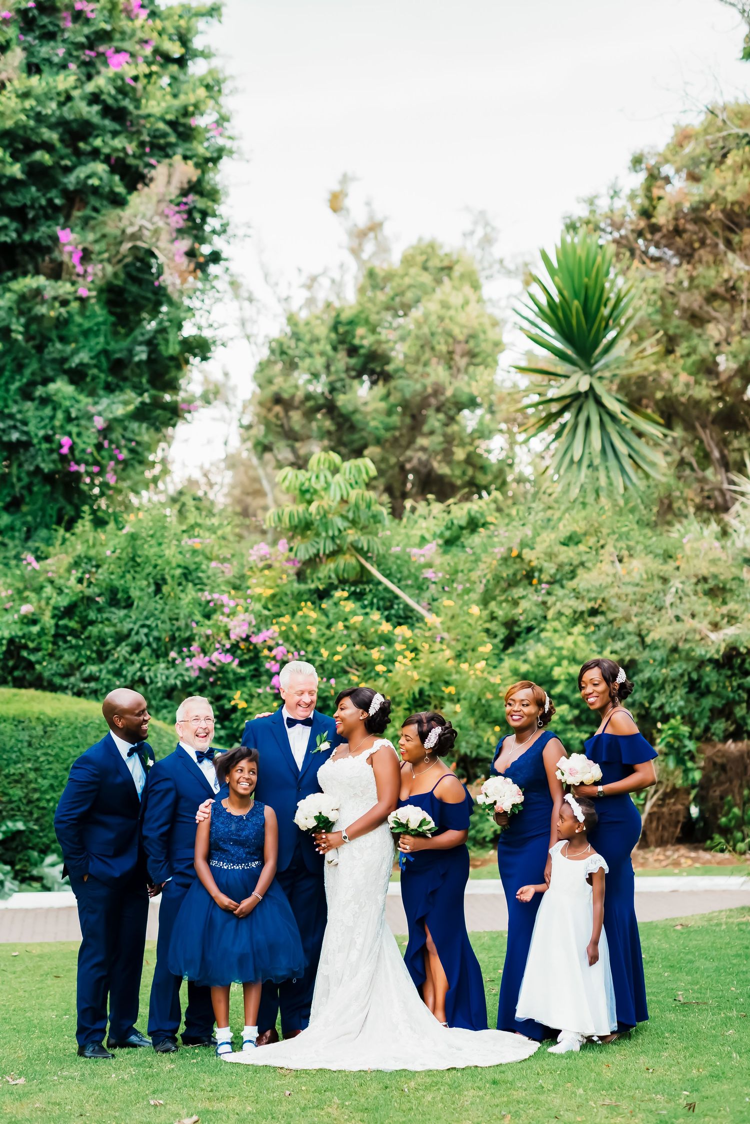 Wedding party in navy blue attire poses playfully on a green lawn with tropical foliage in the background.