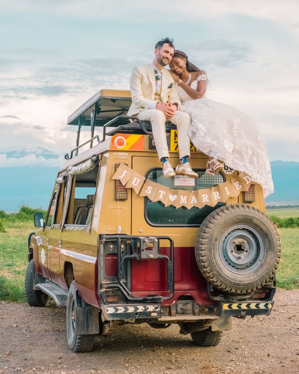 A couple in wedding attire sits atop a vintage yellow safari Land Rover vehicle in an outdoor landscape setting.