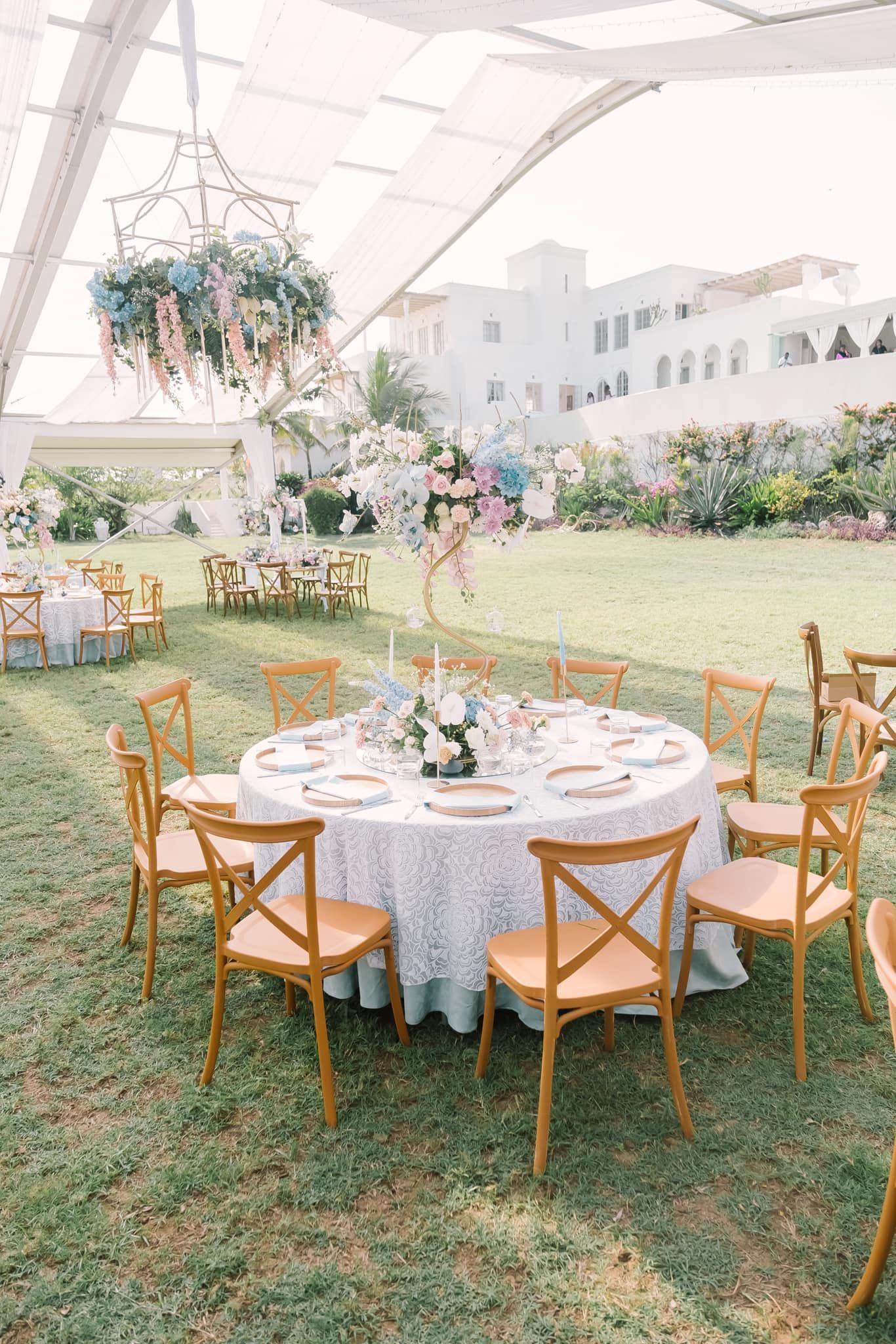 Elegant outdoor wedding reception setup with round tables, white linens and wooden cross-back chairs under a tent canopy.