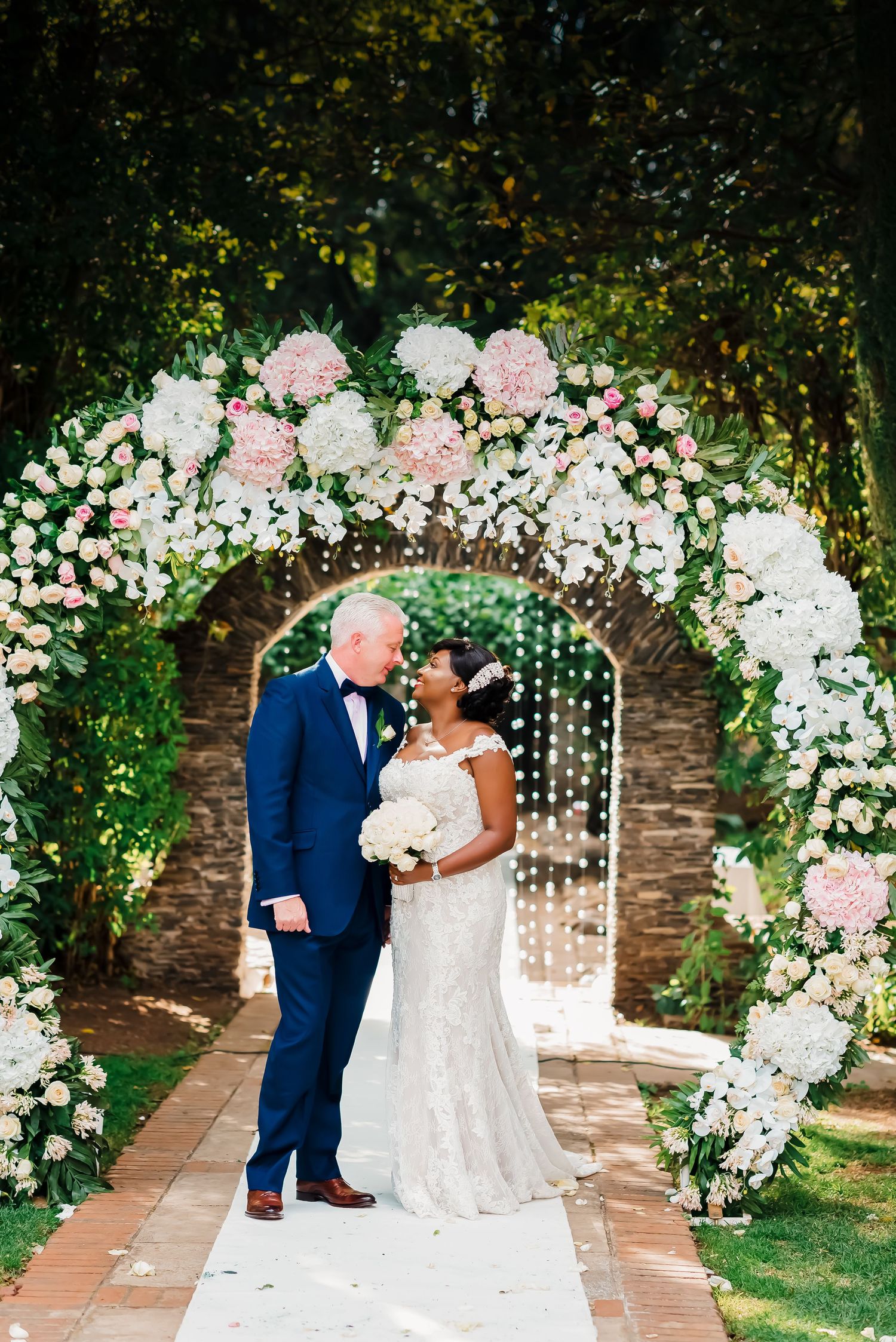 Wedding ceremony under a romantic floral arch with pink and white roses in an outdoor garden setting.
