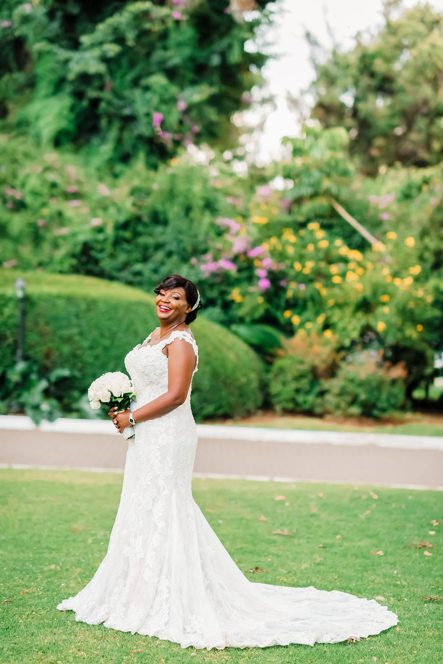 A bride in a flowing white lace wedding gown poses joyfully on a lush green lawn during an outdoor photo session.