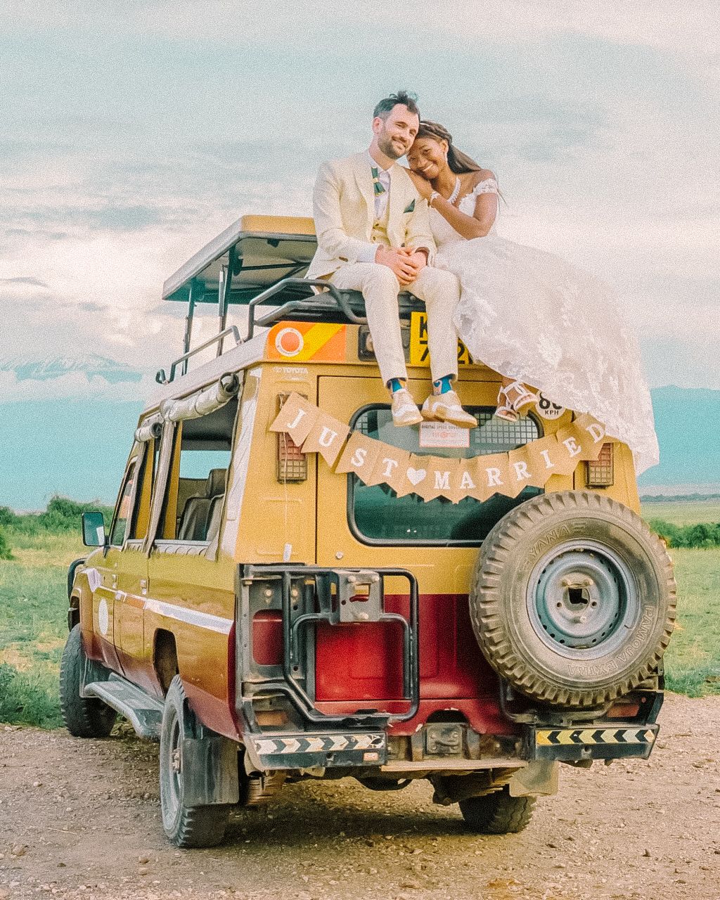 A vintage yellow Land Rover decorated for a wedding with white flowing fabric parked in a scenic outdoor setting.