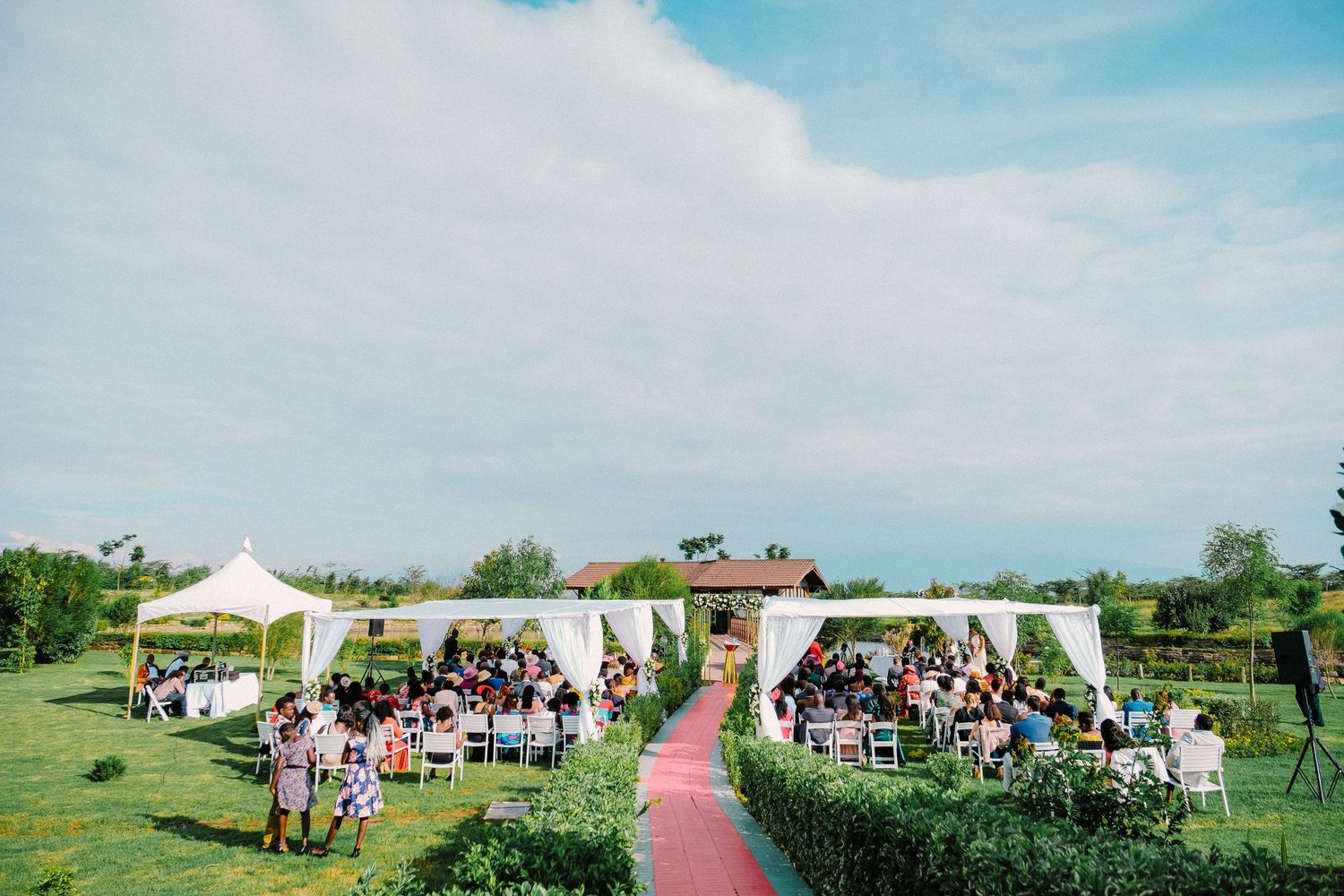Outdoor wedding ceremony setup with white tents, chairs, and a red carpet aisle on a green lawn under blue skies.