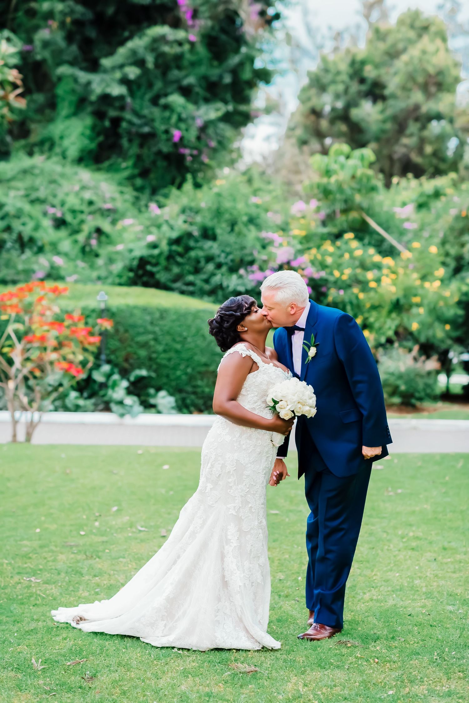 A couple shares a romantic kiss in a lush garden setting during a wedding celebration.
