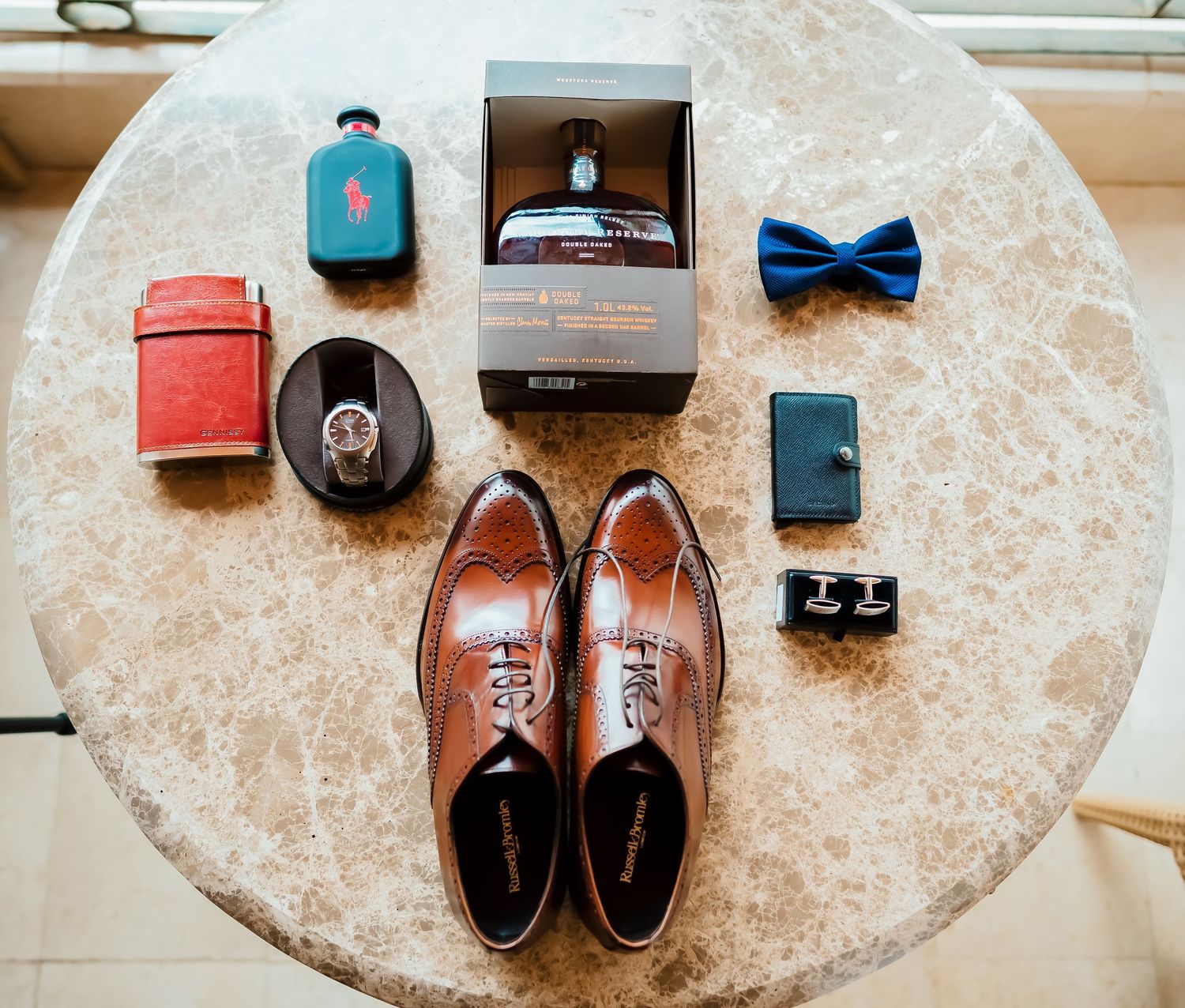 Brown leather dress shoes and accessories including bow tie and cufflinks arranged on marble surface.