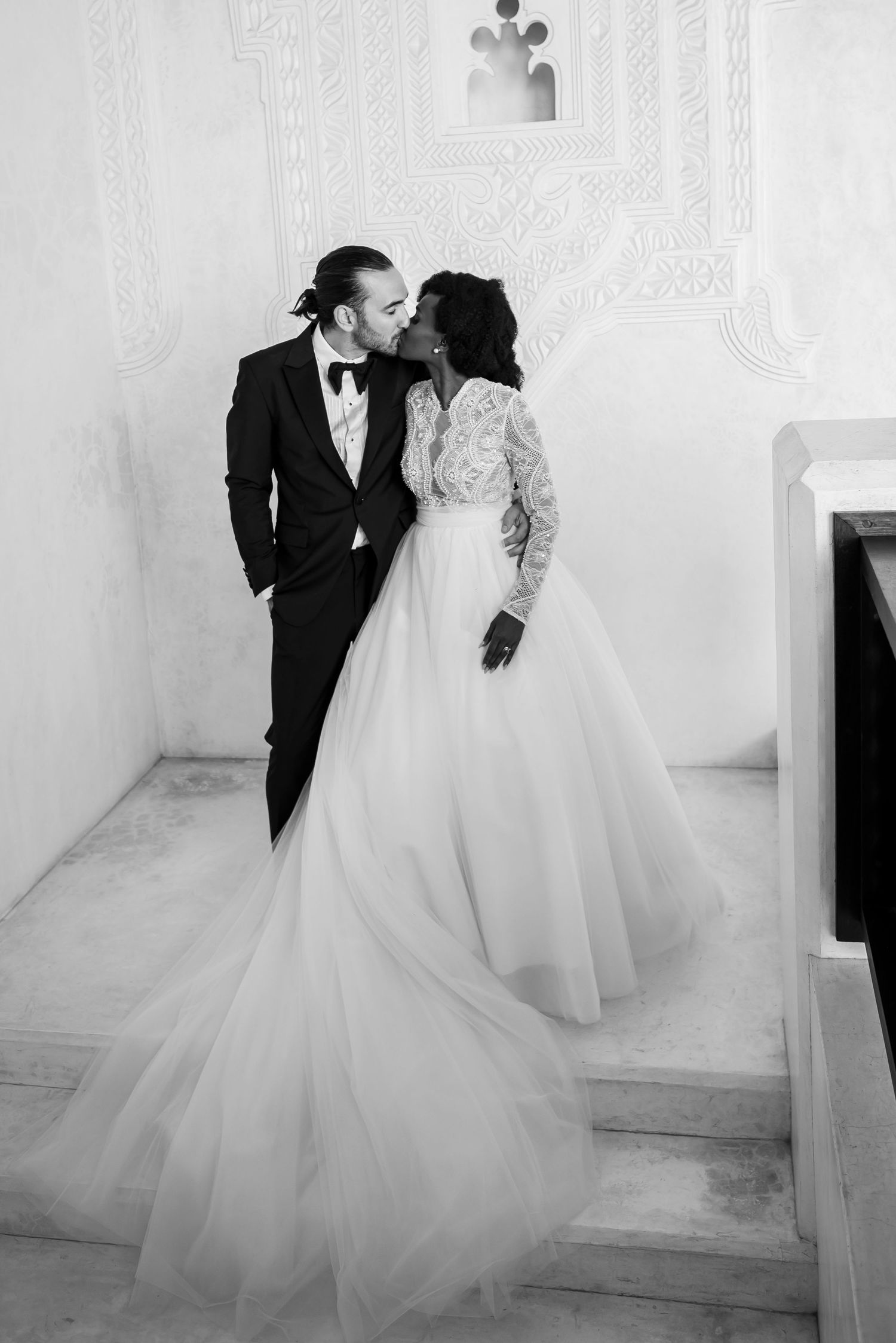 Black and white wedding photo series showing romantic moments between couple in elegant dress and tuxedo against white wall.