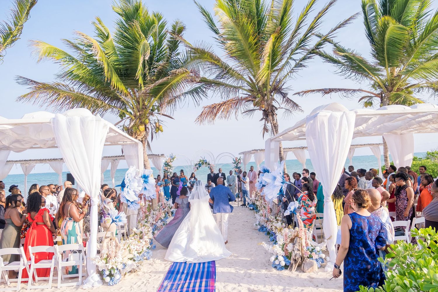 A scenic beach wedding ceremony setup with white chairs and palm trees along the shoreline under bright blue skies.