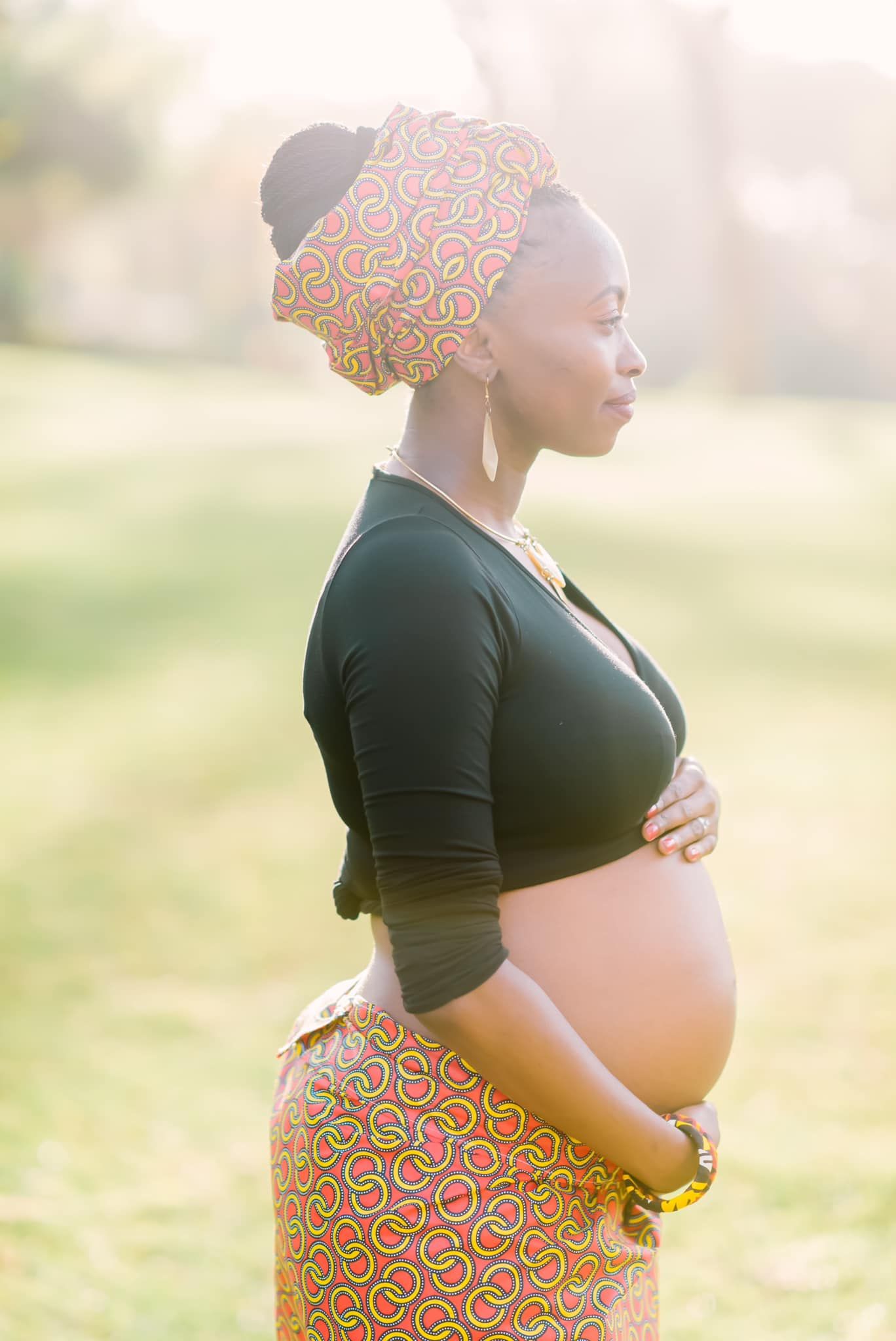 A pregnant figure in traditional African print skirt and black top stands in soft sunlit setting during maternity photoshoot.