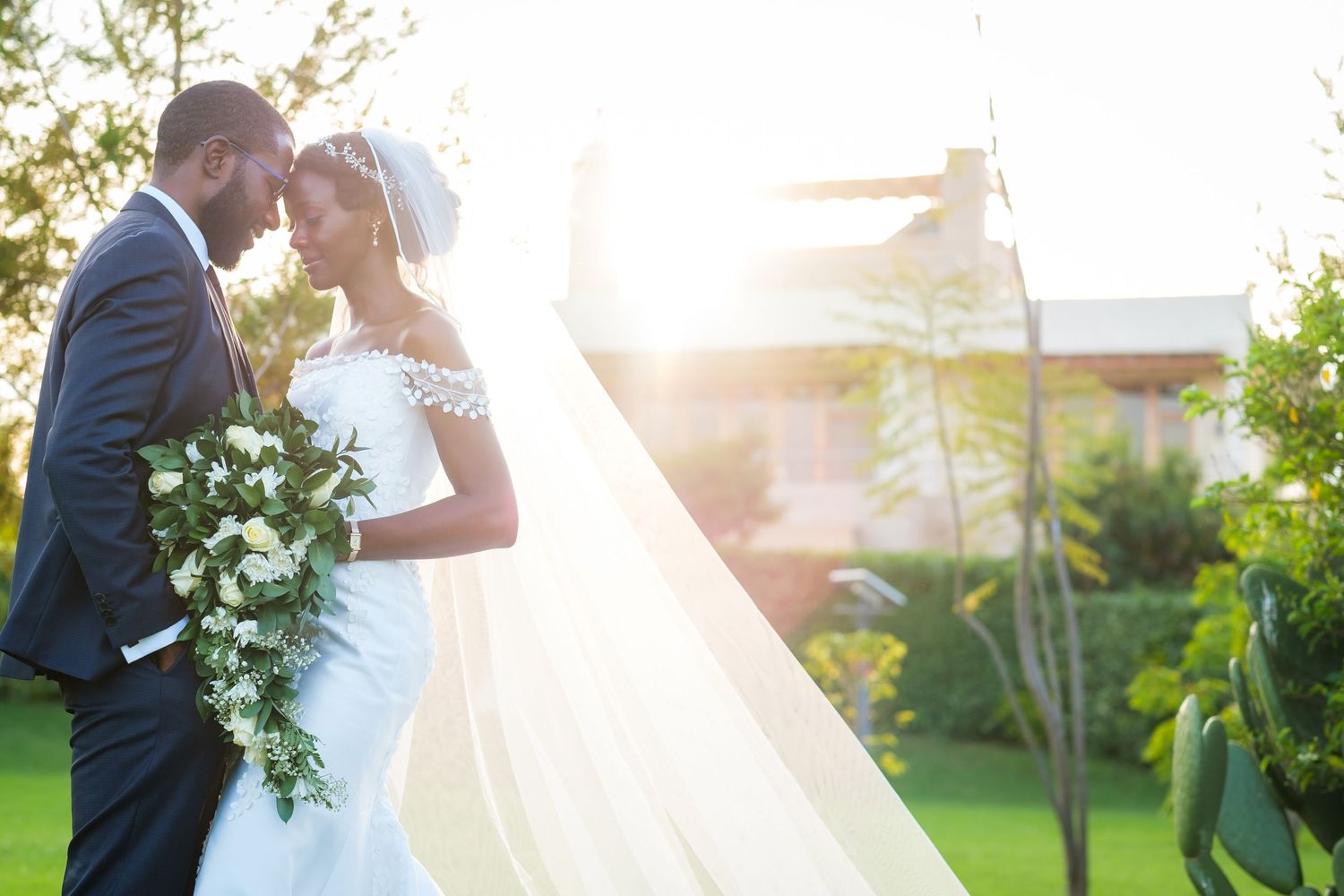 A couple shares an intimate moment in a sunlit garden setting on their wedding day, with a flowing white veil and floral bouquet.