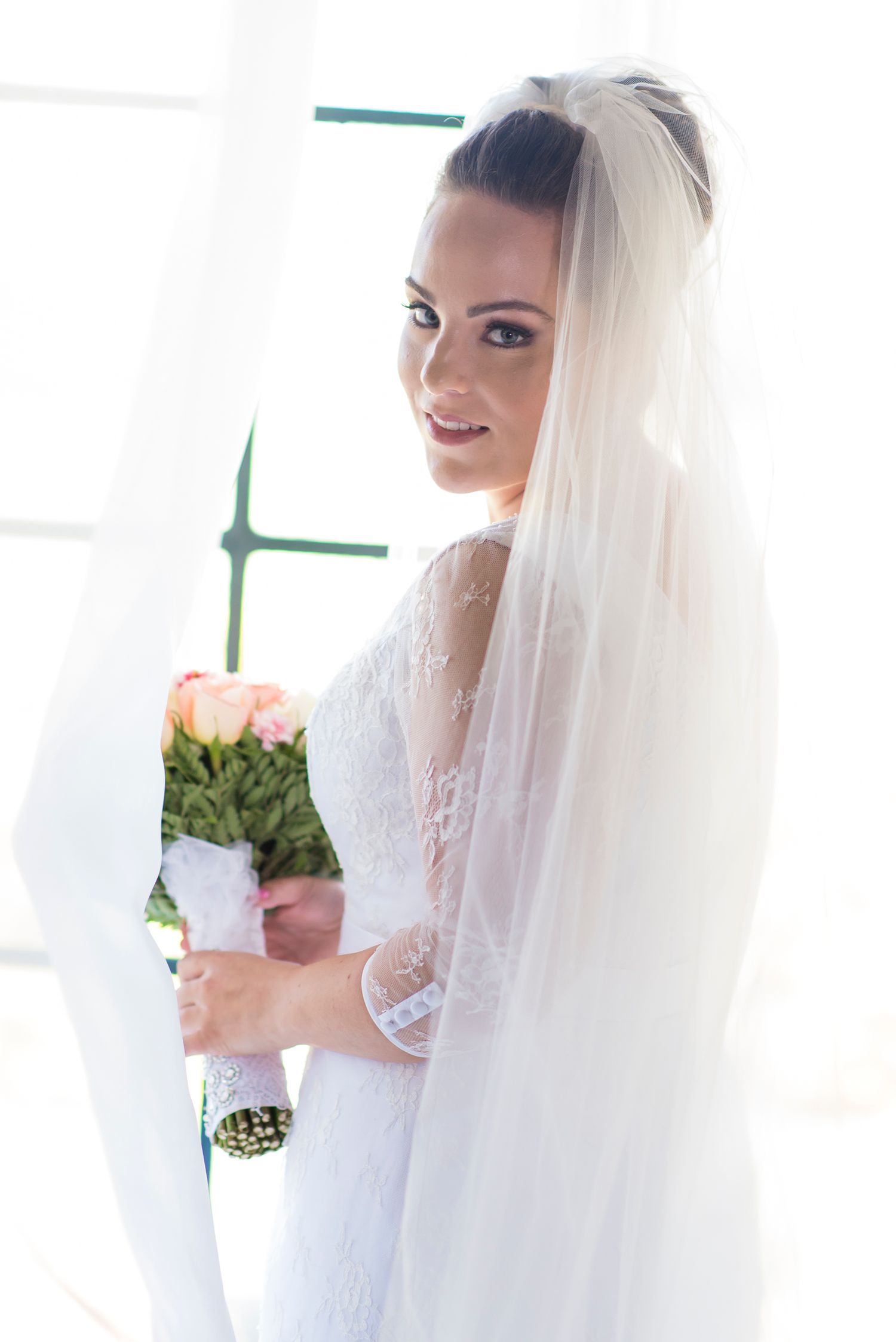 A bride in a white lace wedding dress and long veil holds a bouquet of pink roses near a bright window.
