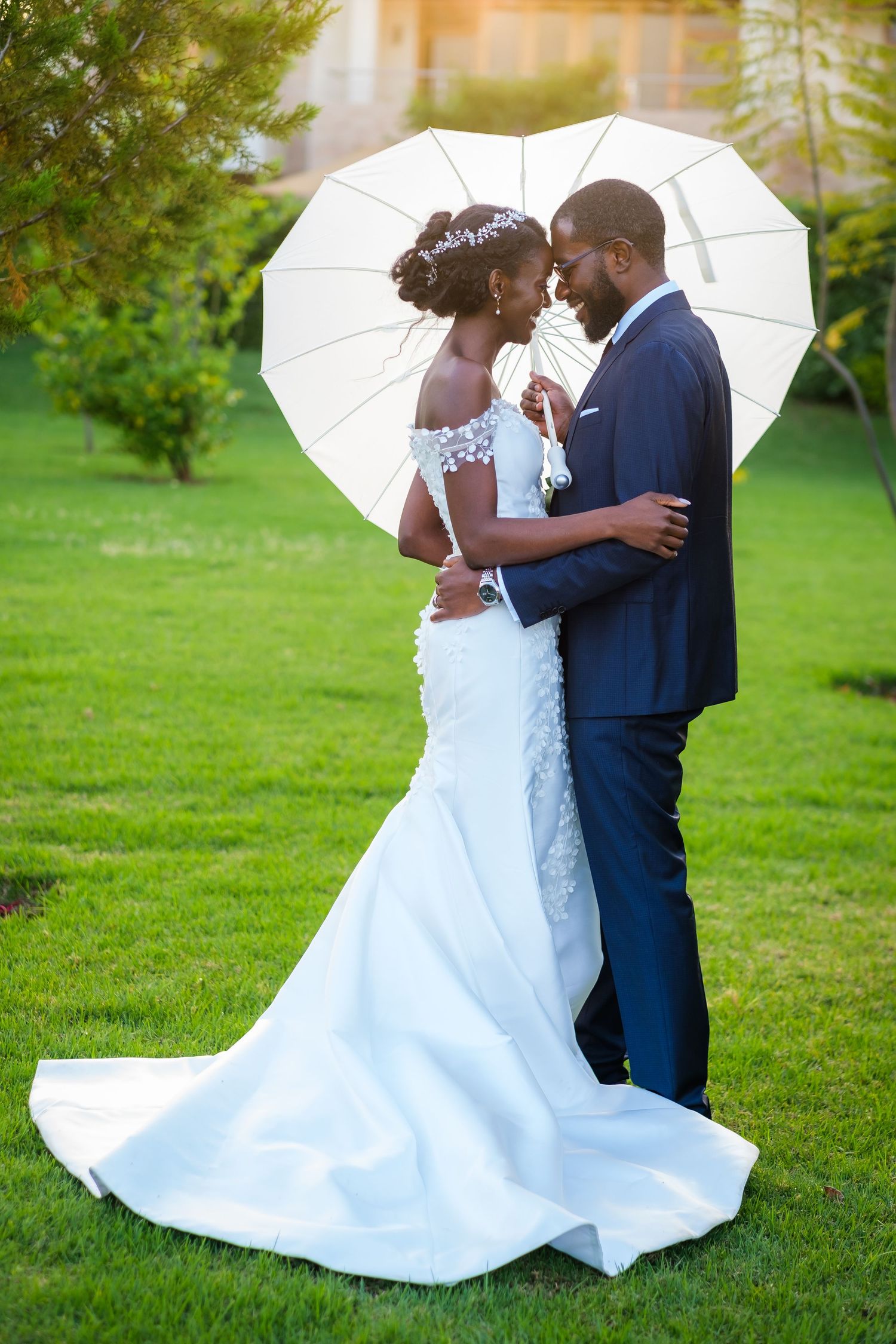 A couple shares an intimate moment under a white umbrella on their wedding day in a lush green garden setting.