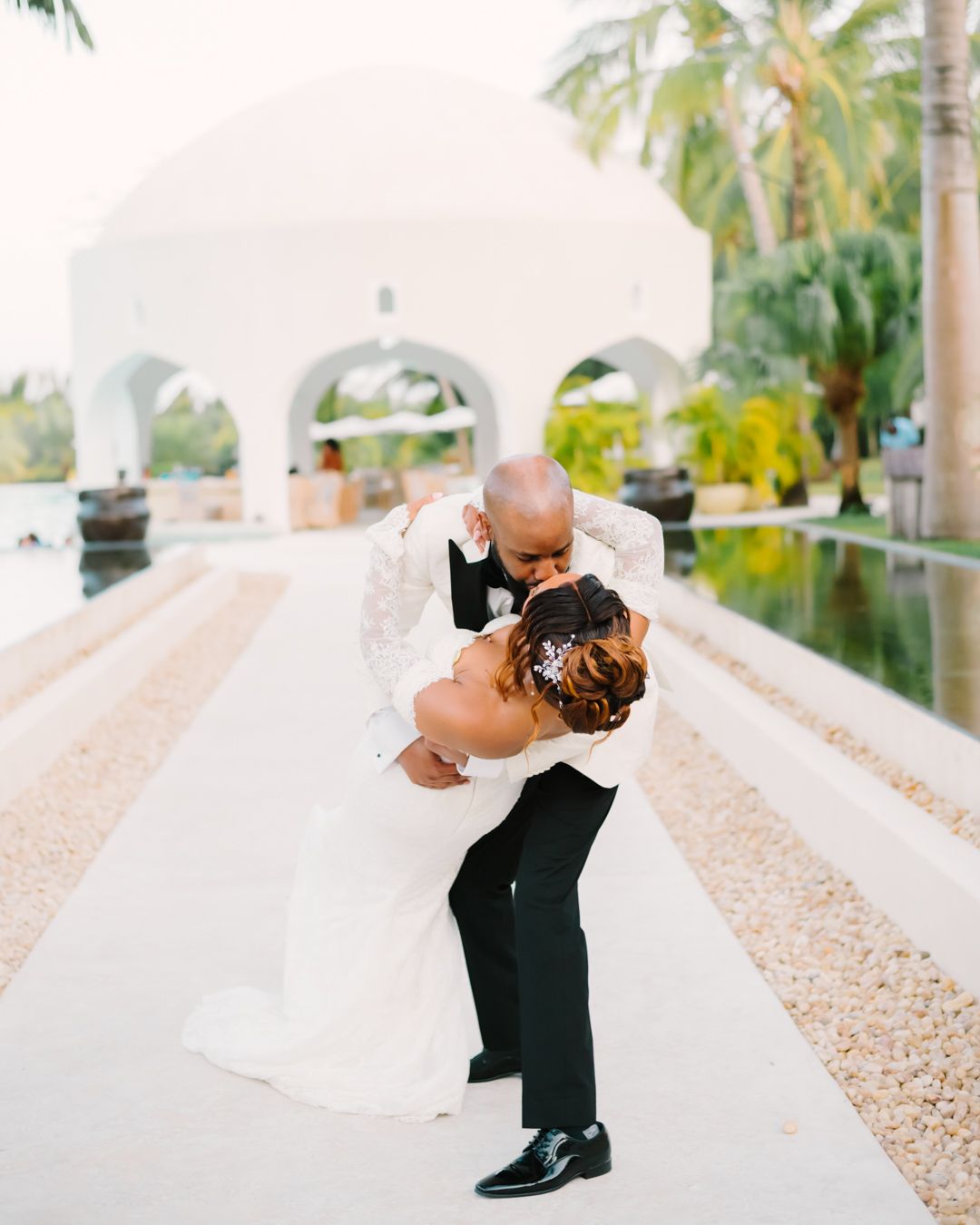 A romantic wedding couple embraces and kisses in a white archway with palm trees in the background.