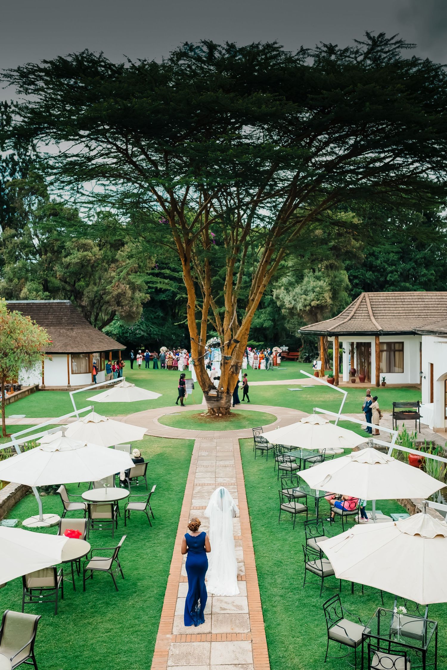 Panoramic view of an outdoor event venue with white canopies, brick pathways, and lush trees in a manicured garden setting.