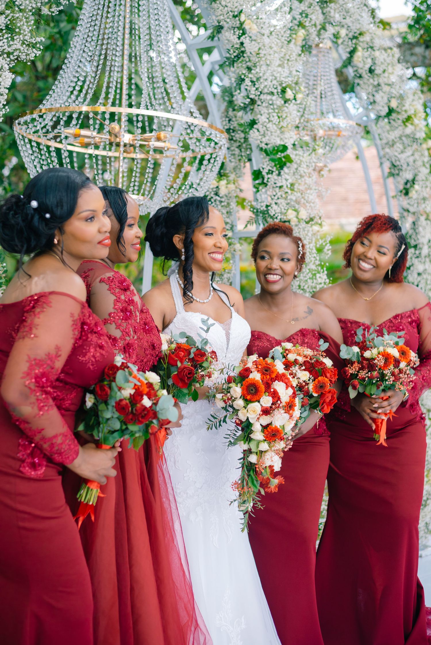 Wedding party in burgundy dresses and white gowns gather under floral decorations and crystal chandeliers.