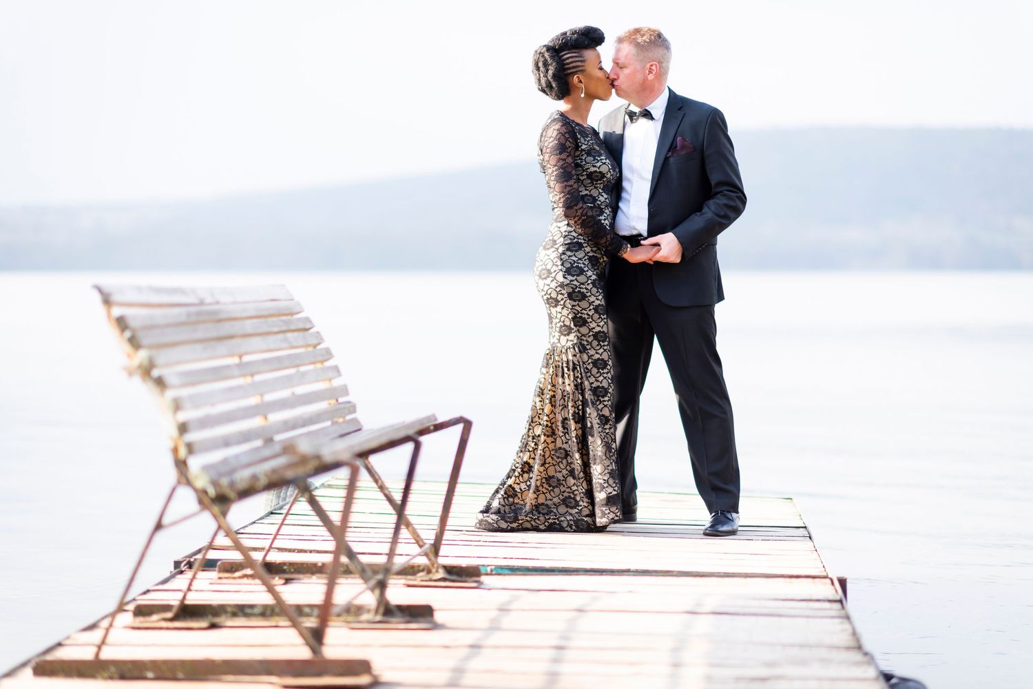 Couple in formal wedding attire embraces on a snowy winter beach with wooden beach chairs in the background.
