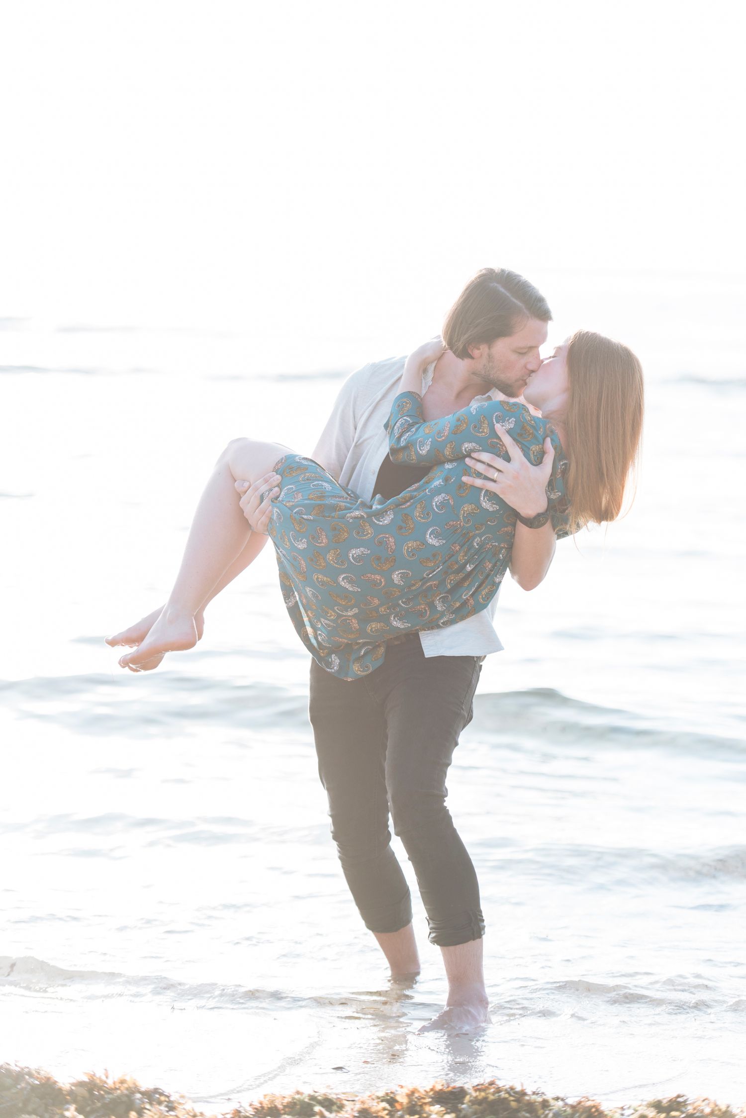 Couple playfully splashing and embracing at the beach during sunset with waves rolling in behind them.