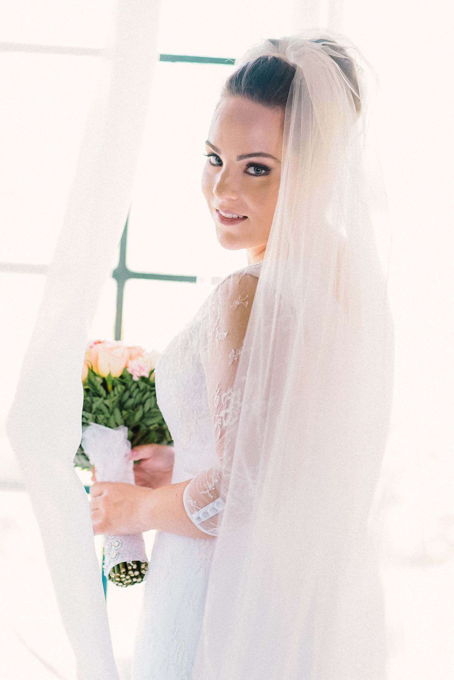 Bride in white lace wedding dress and veil holds delicate pink rose bouquet in bright sunlit room.