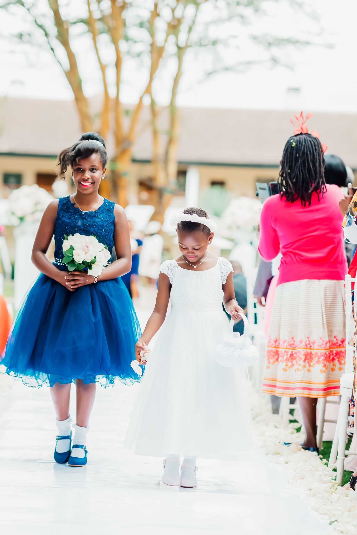 Two attendants in formal dresses walk down an outdoor path during a wedding ceremony.