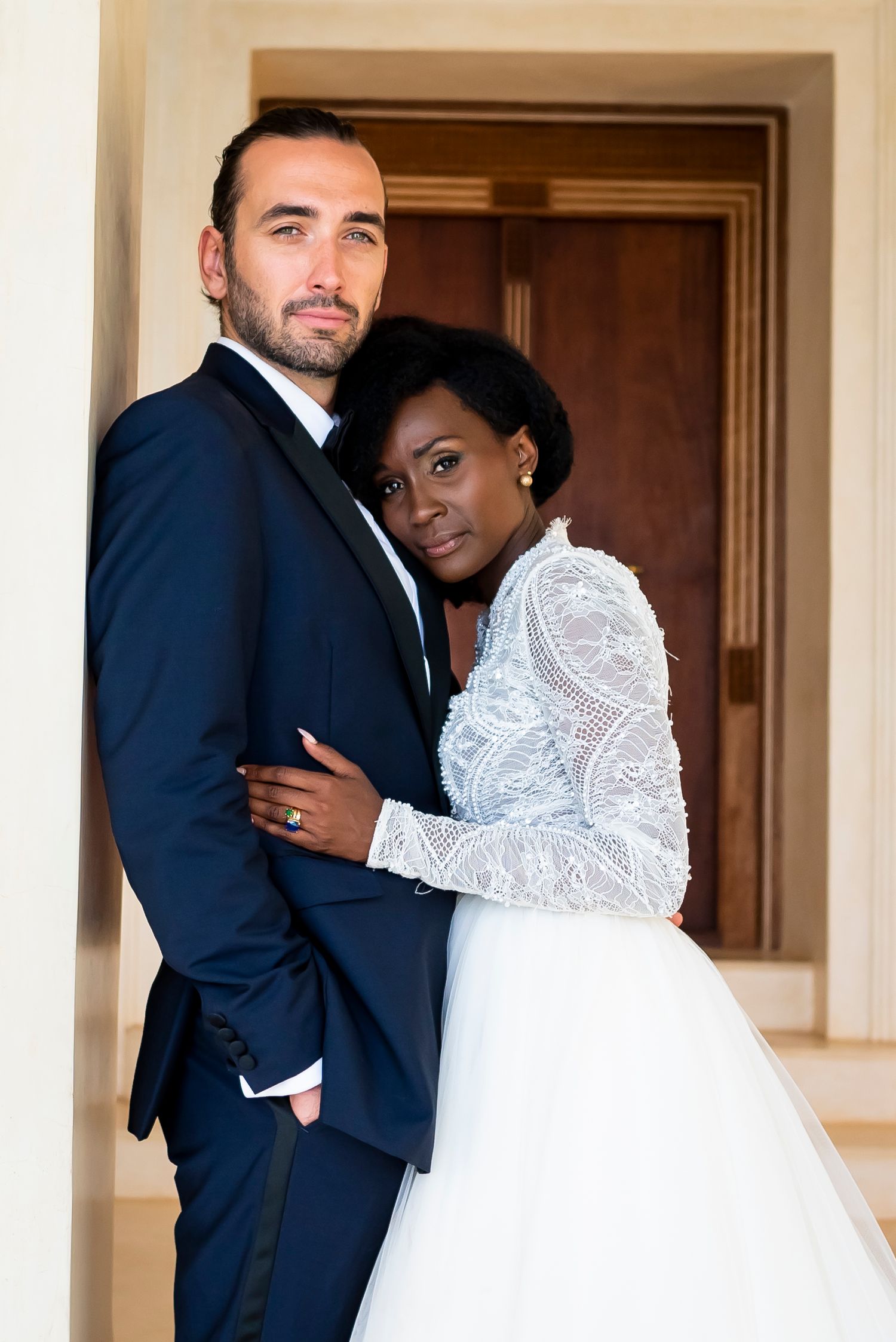 Elegant interracial couple in wedding attire pose together in a doorway, bride wearing lace white gown.