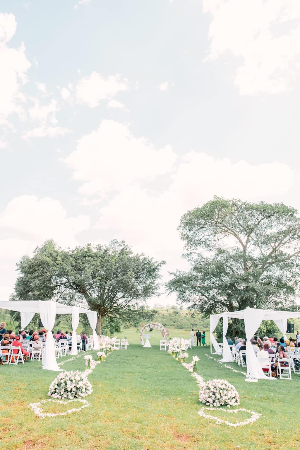 Elegant outdoor wedding ceremony setup with two white gazebos, floral arrangements, and white chairs on a green lawn.