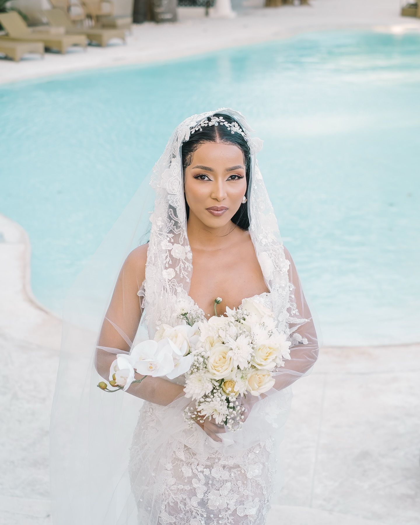 A bride in a lace wedding dress poses elegantly by a serene pool surrounded by tropical landscaping.
