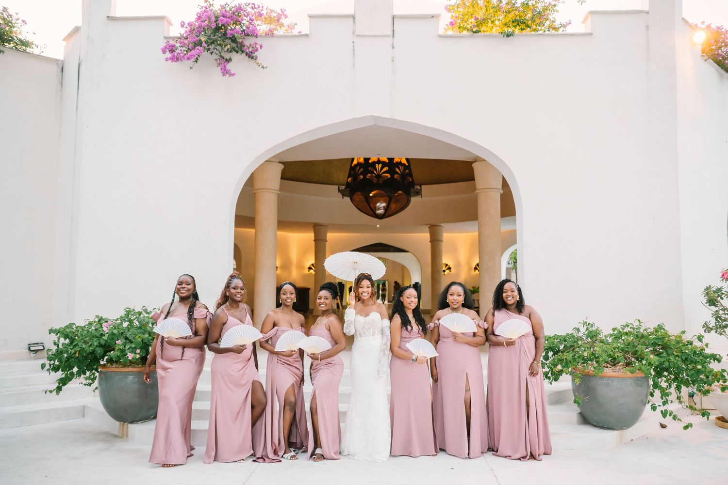 Wedding party in pink bridesmaid dresses and white bridal gown pose under Mediterranean-style white archway with flowers.