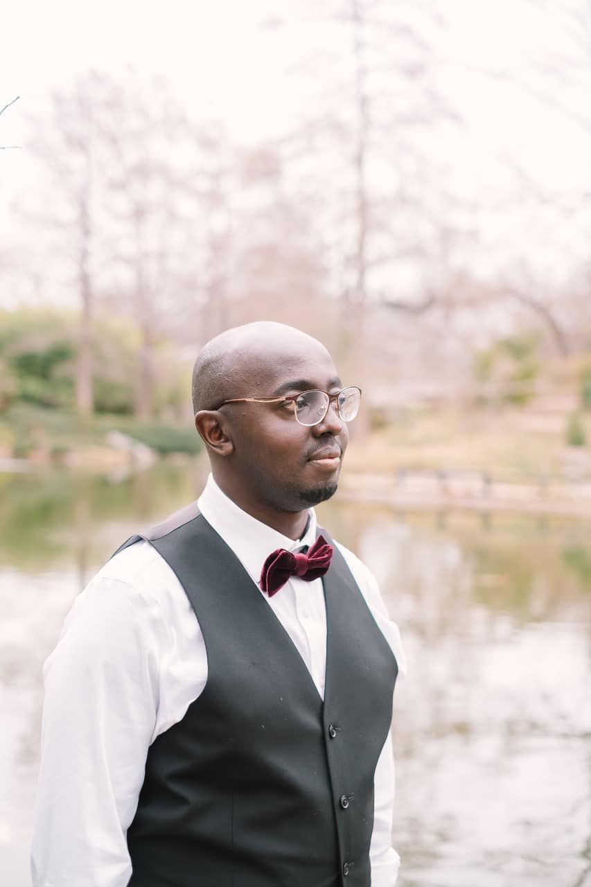 A person in formal attire with a burgundy bow tie and black vest stands outdoors against a soft, blurred nature backdrop.