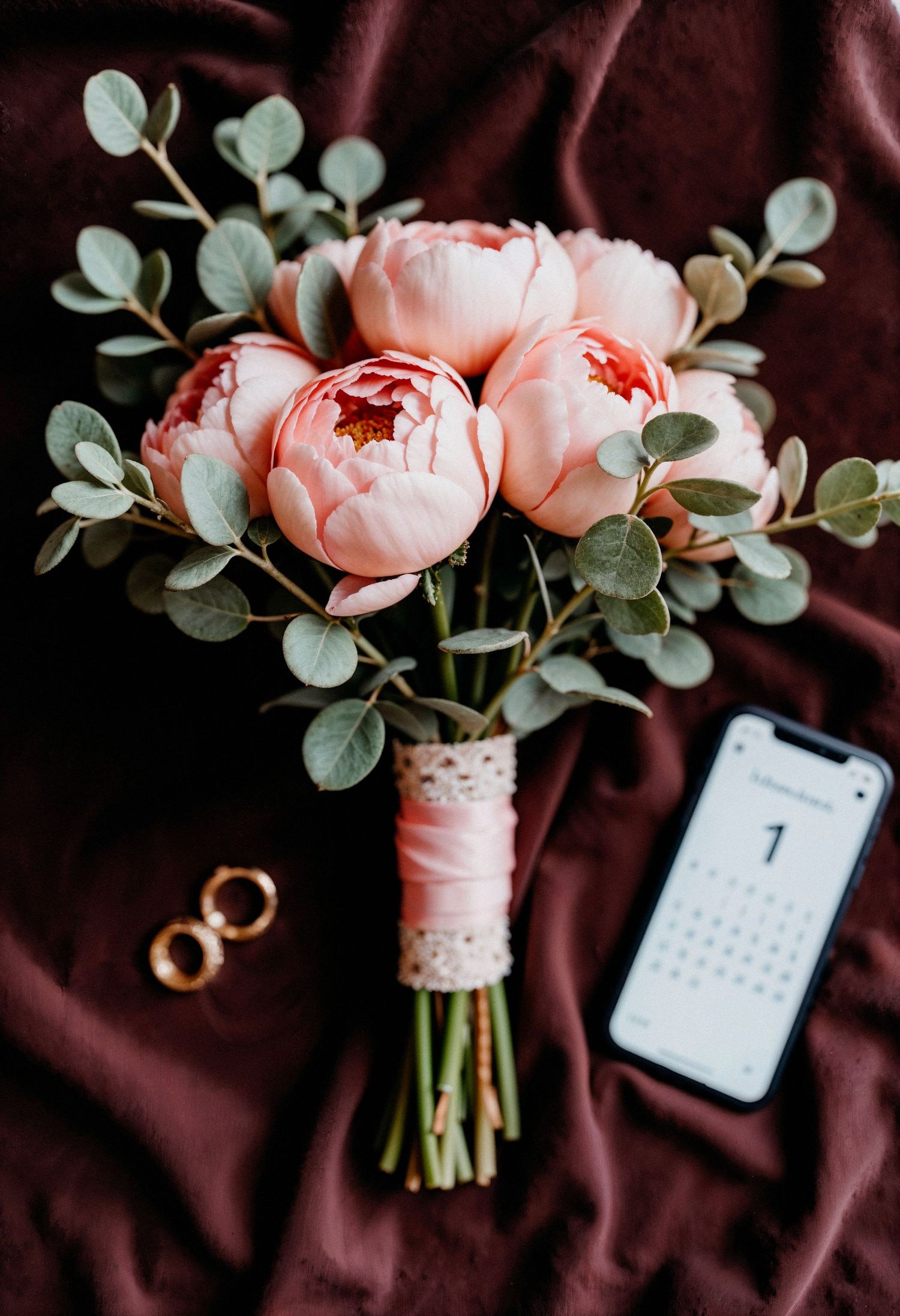 A romantic arrangement of pink peonies with eucalyptus leaves against a dark burgundy backdrop with a calendar and ring.
