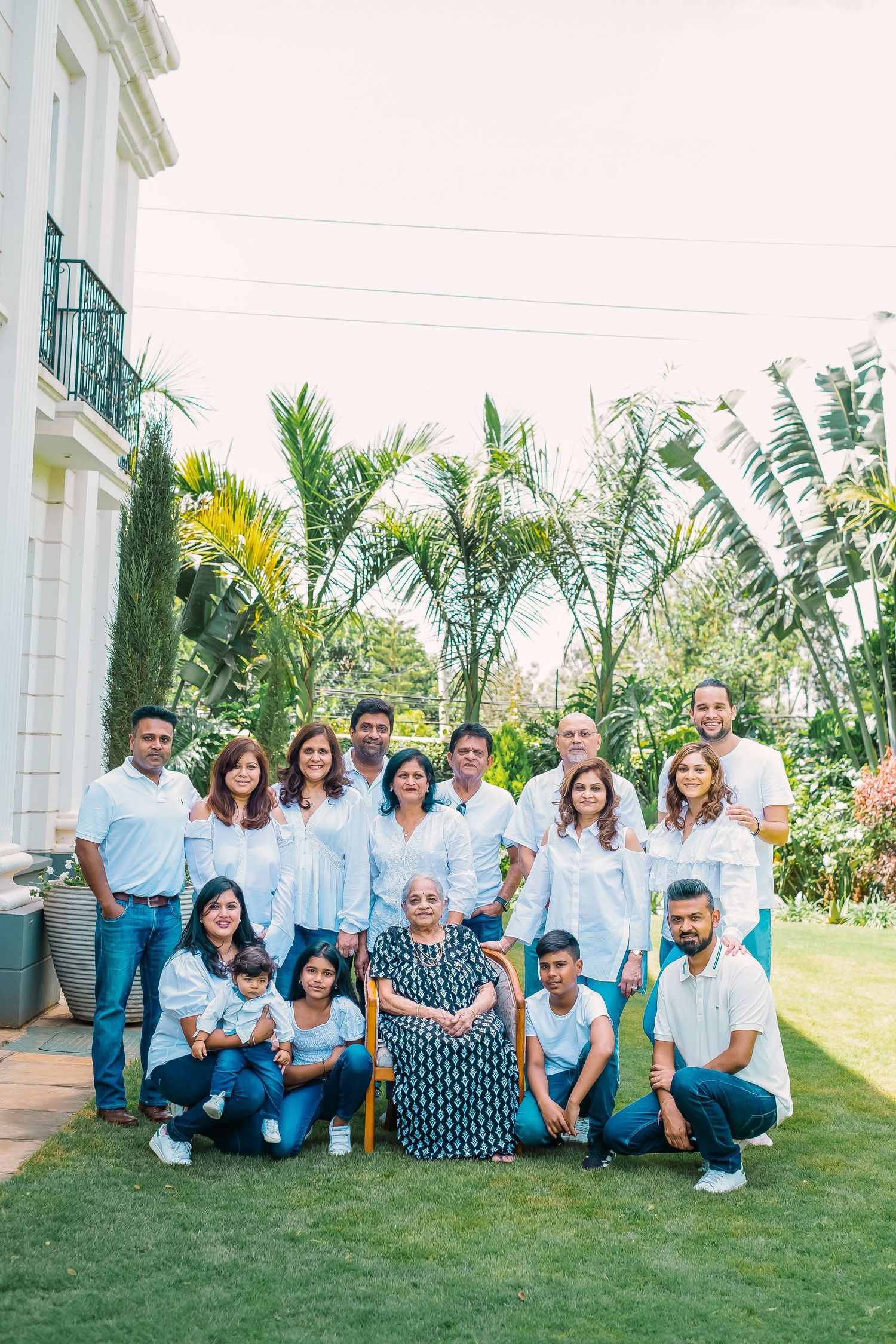Large family group sitting together on grass lawn with palm trees and white building in background.