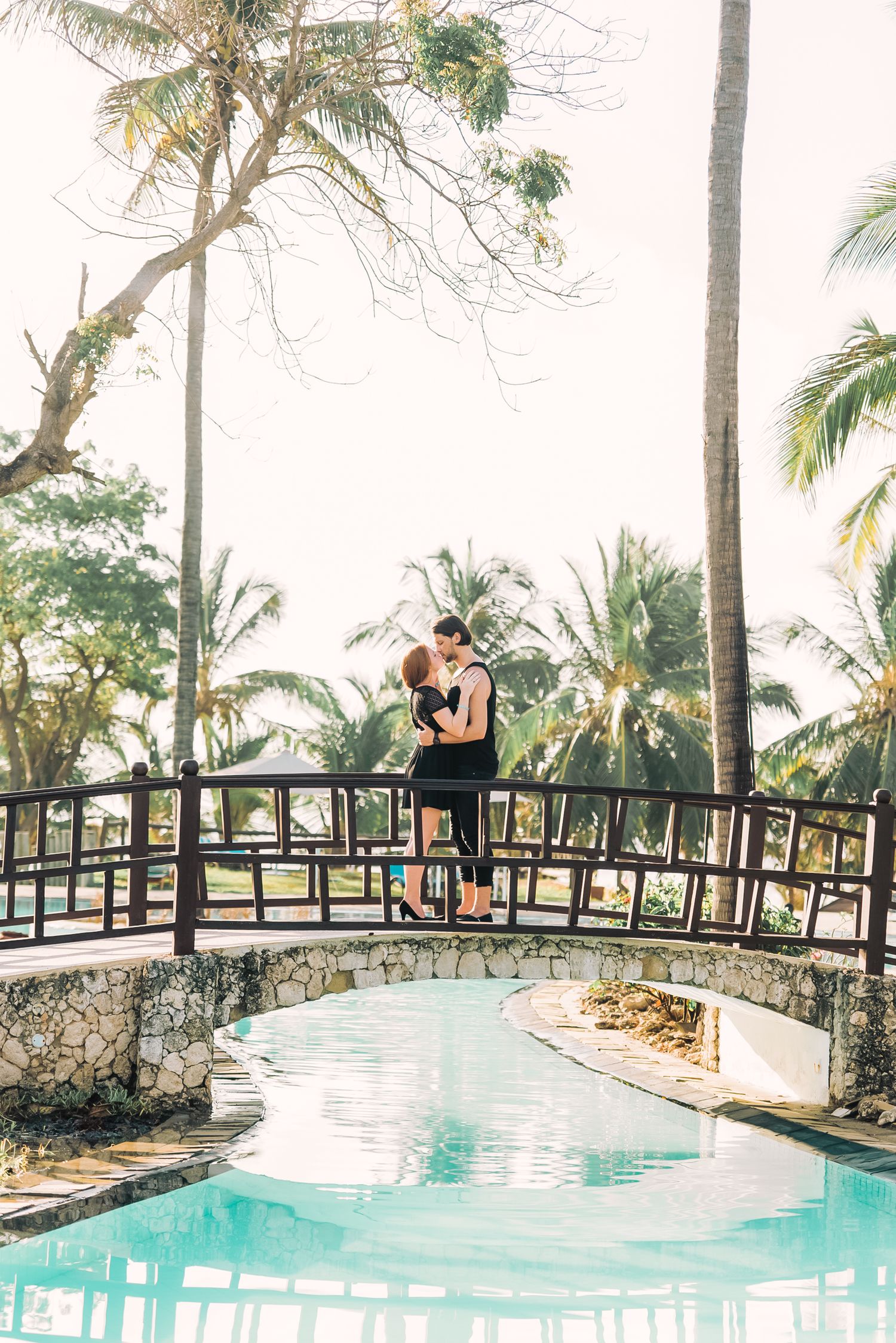 A couple embraces on a curved wooden bridge over turquoise water surrounded by tropical palm trees at a resort.